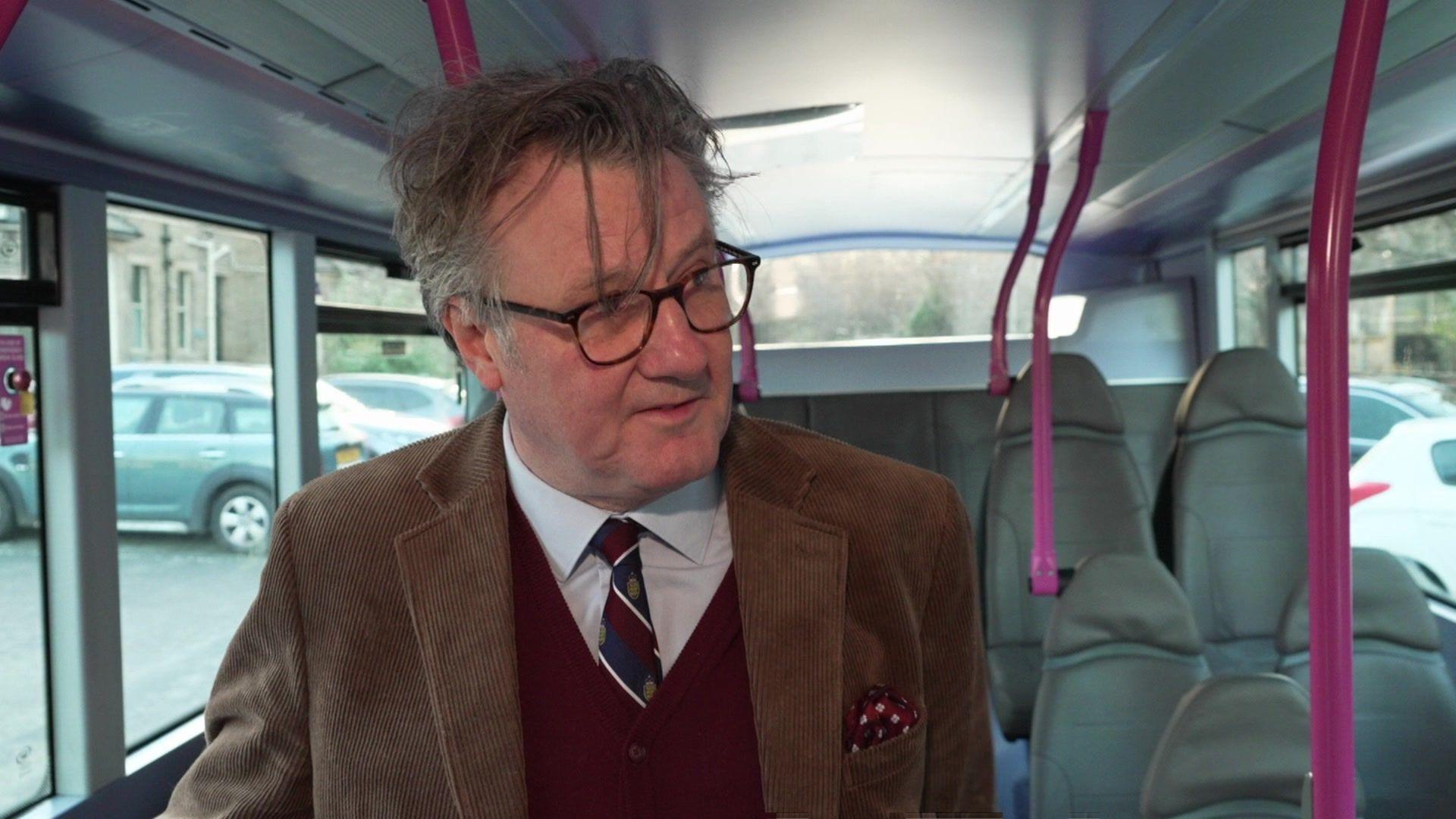 A man wearing glasses and in a brown jacket, mauve pullover and a shirt and tie, stands inside a bus.