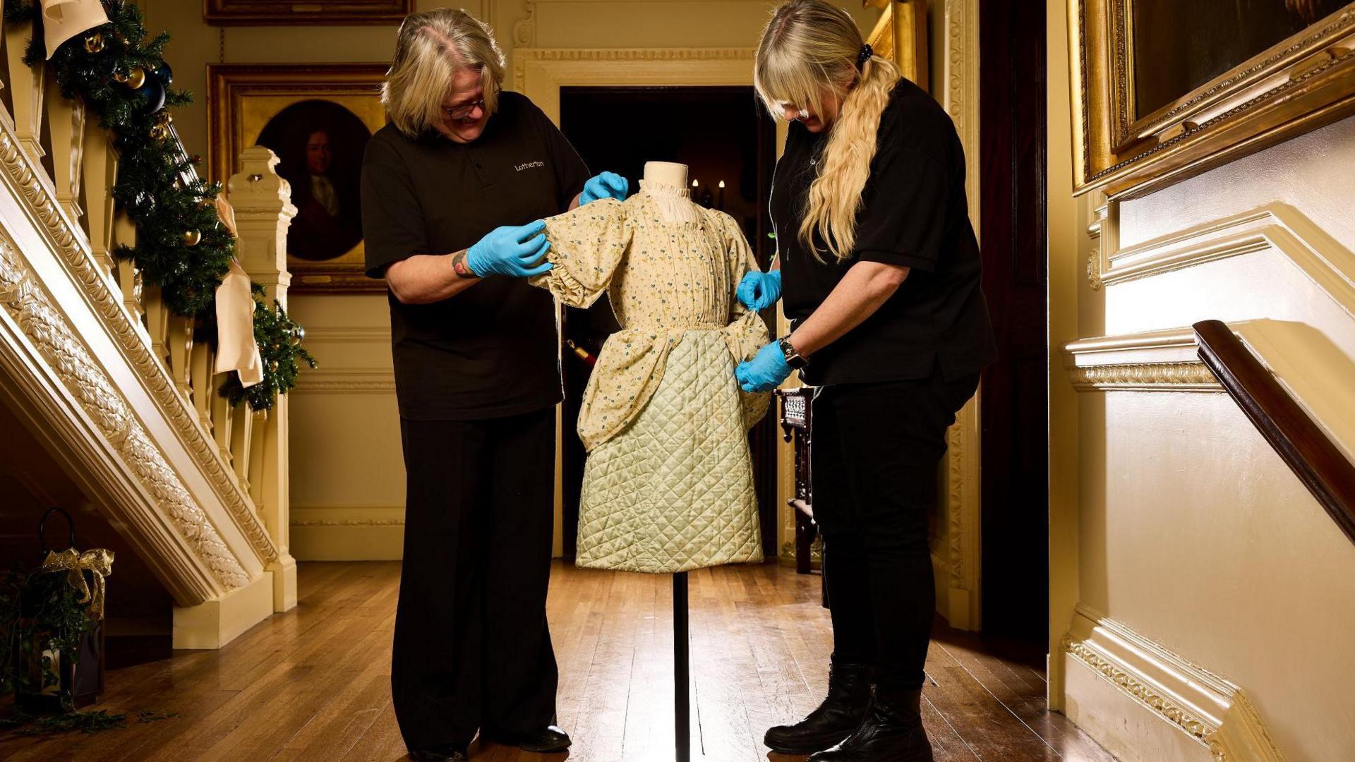 Two women dressed in black tending to a child's dress on a clothes stand. They are stood in the hall of a grand house.