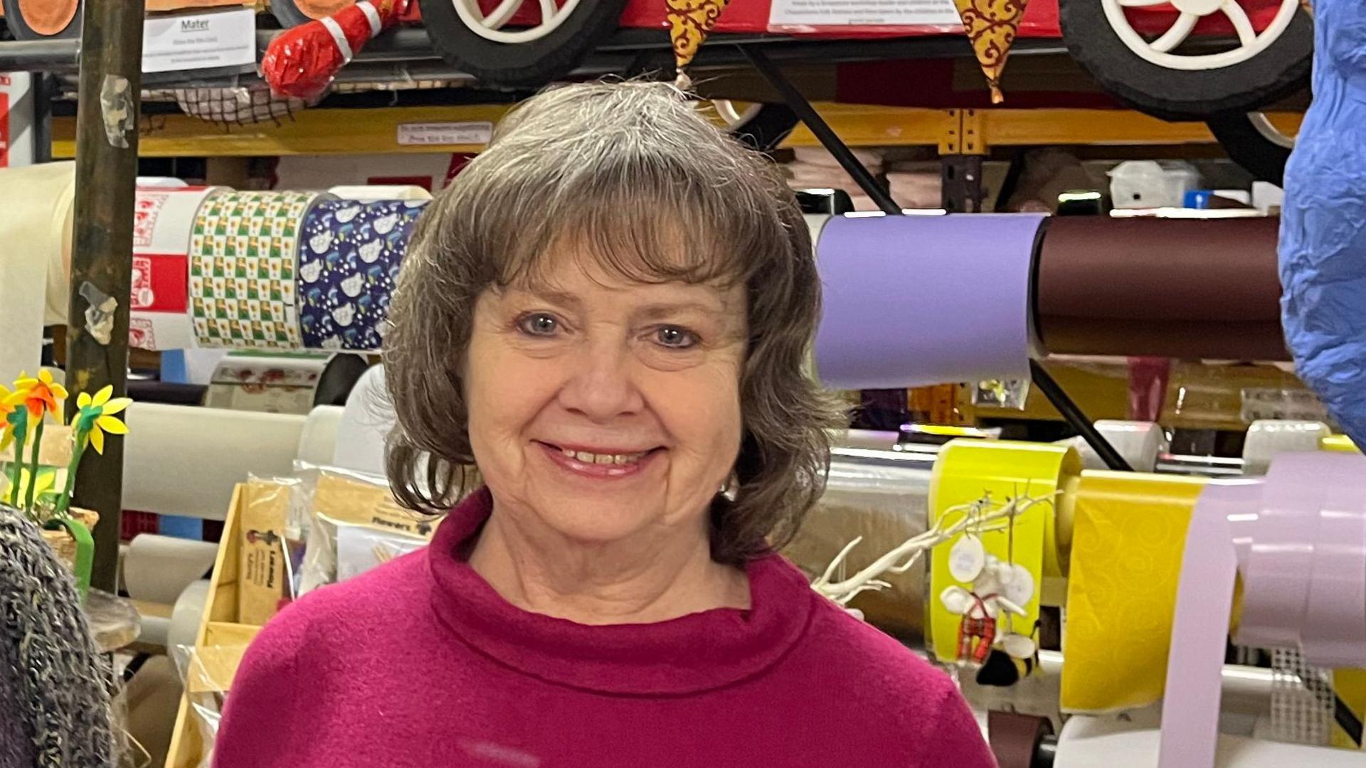 A woman wearing a pink jumper, smiles at the camera while standing in a warehouse with rolls of wrapping paper behind her. The wheels of red toy a car can be partially seen above her head. She has a brown/grey bob and fringe.