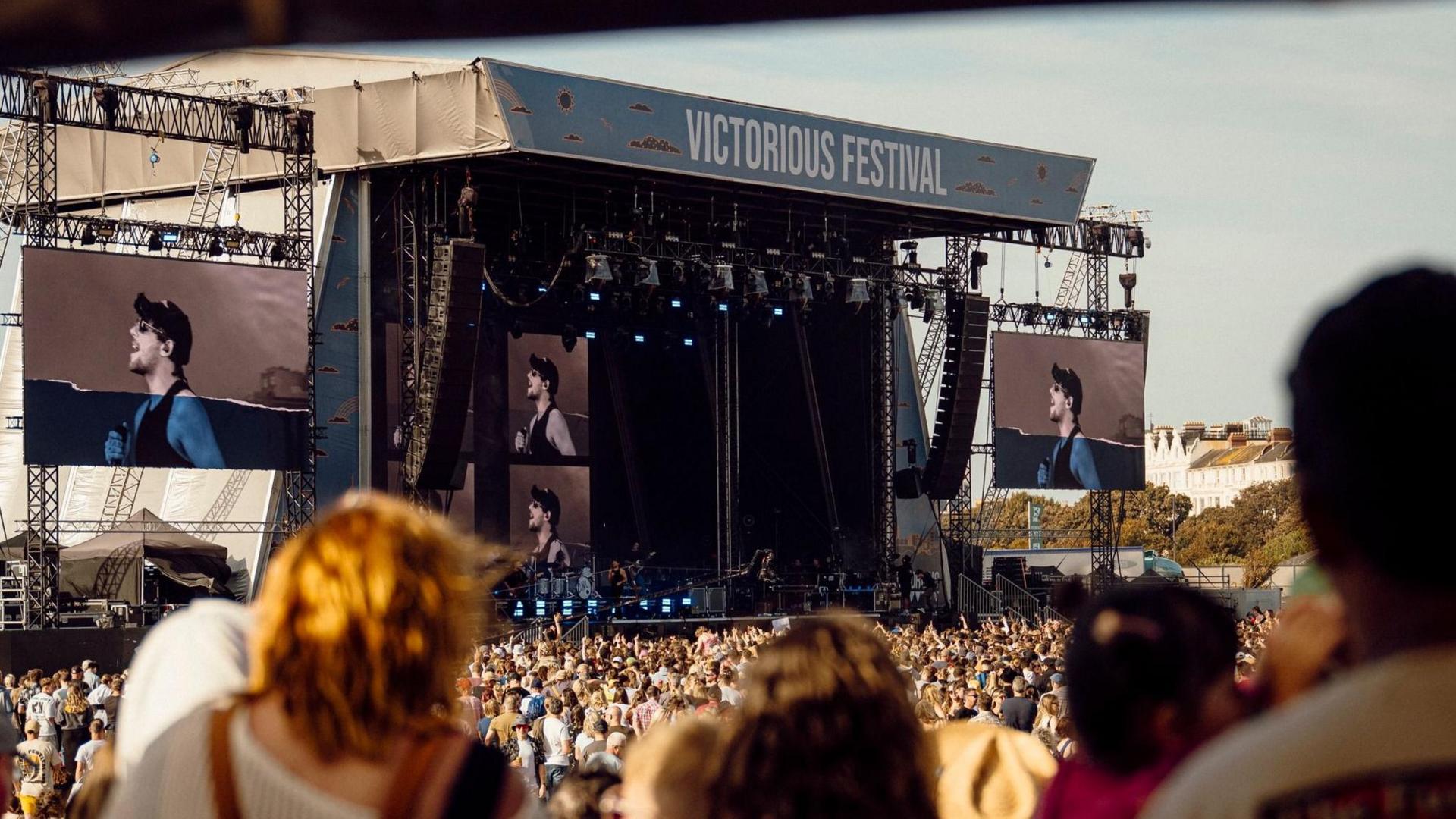 Stage in the background with large crown in front and the backs of people's heads as they watch a music act.