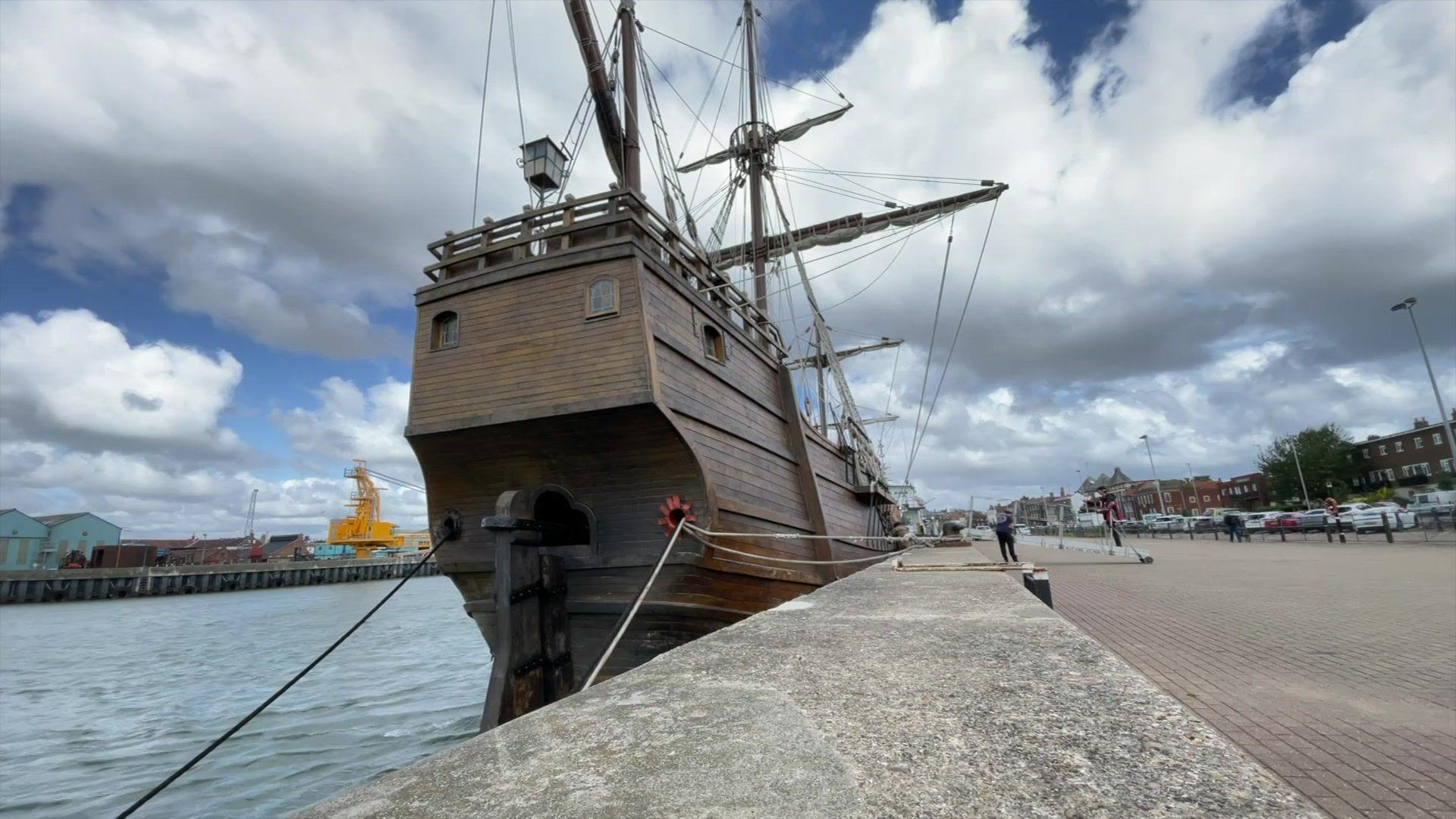 An image of the ship from its rear. Long ropes secure the ship in place in the harbour.