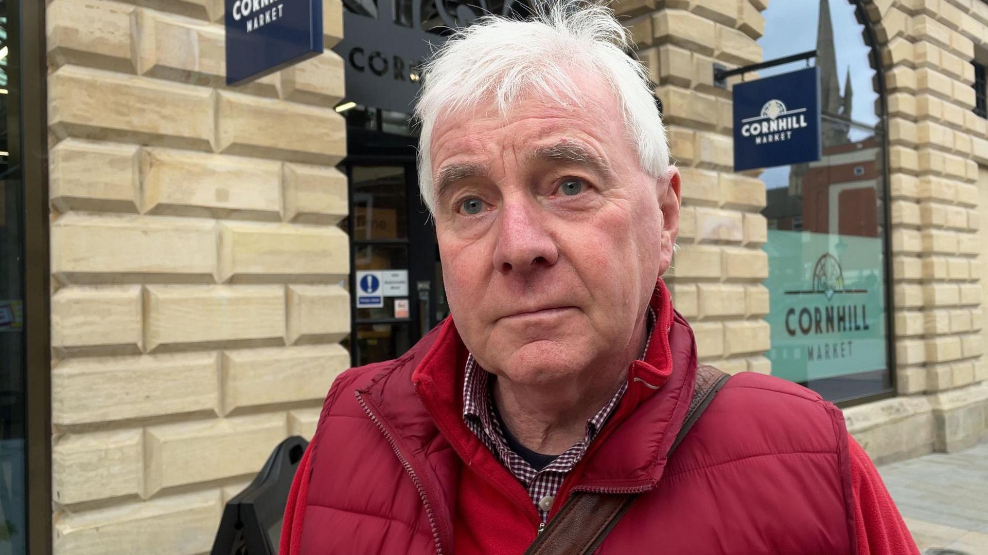 A grey haired man wearing a red gilet over a red fleece looks at the camera. He is standing in front of a large, lightly-coloured stone building which has large windows. There is a sign which reads Cornhill Market.