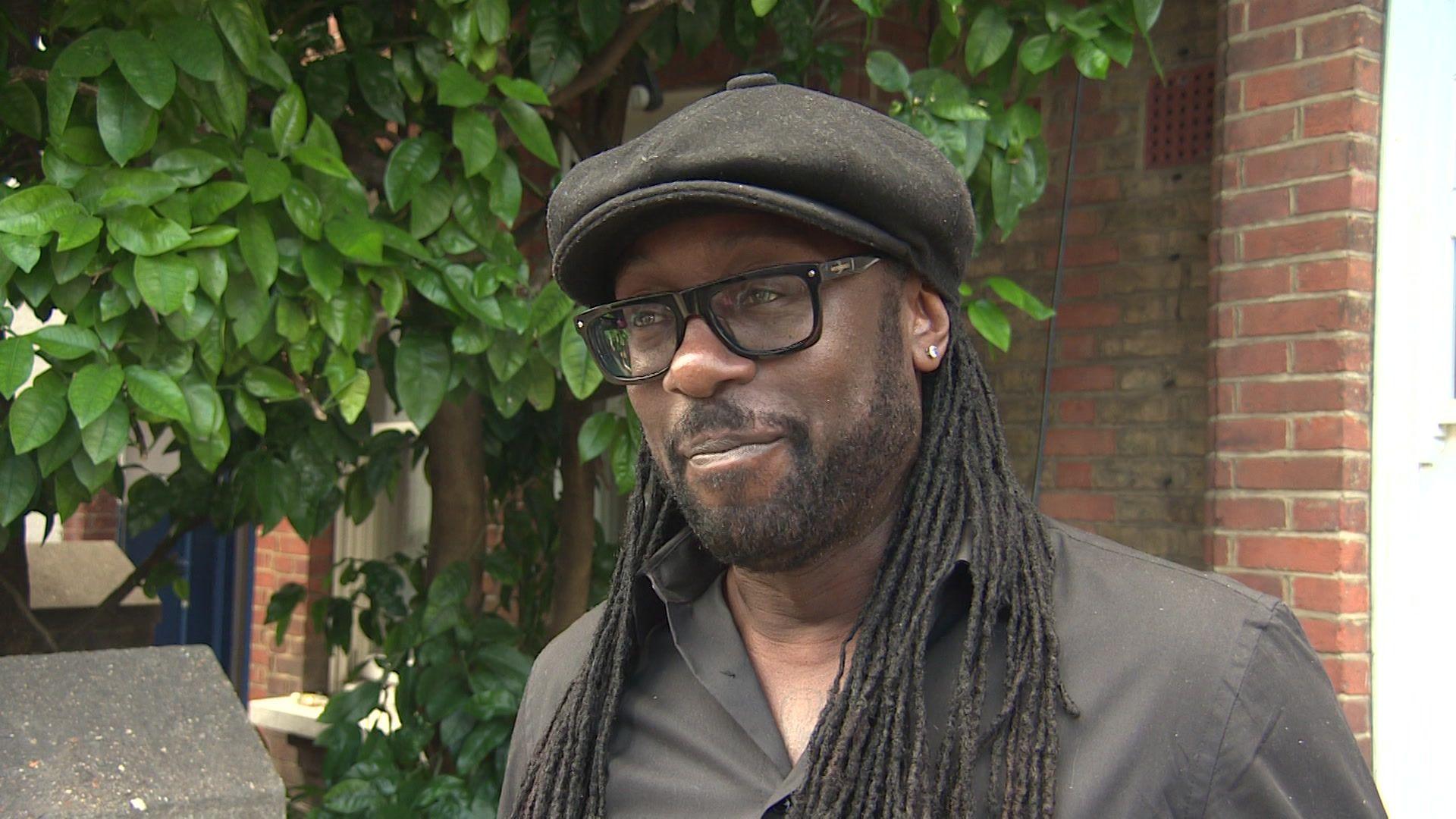 Paul Sealy, a man with long dreadlocks, wearing a large black flat cap hat, black framed glasses and a black shirt, he is standing outside his house next to the grapefruit tree 