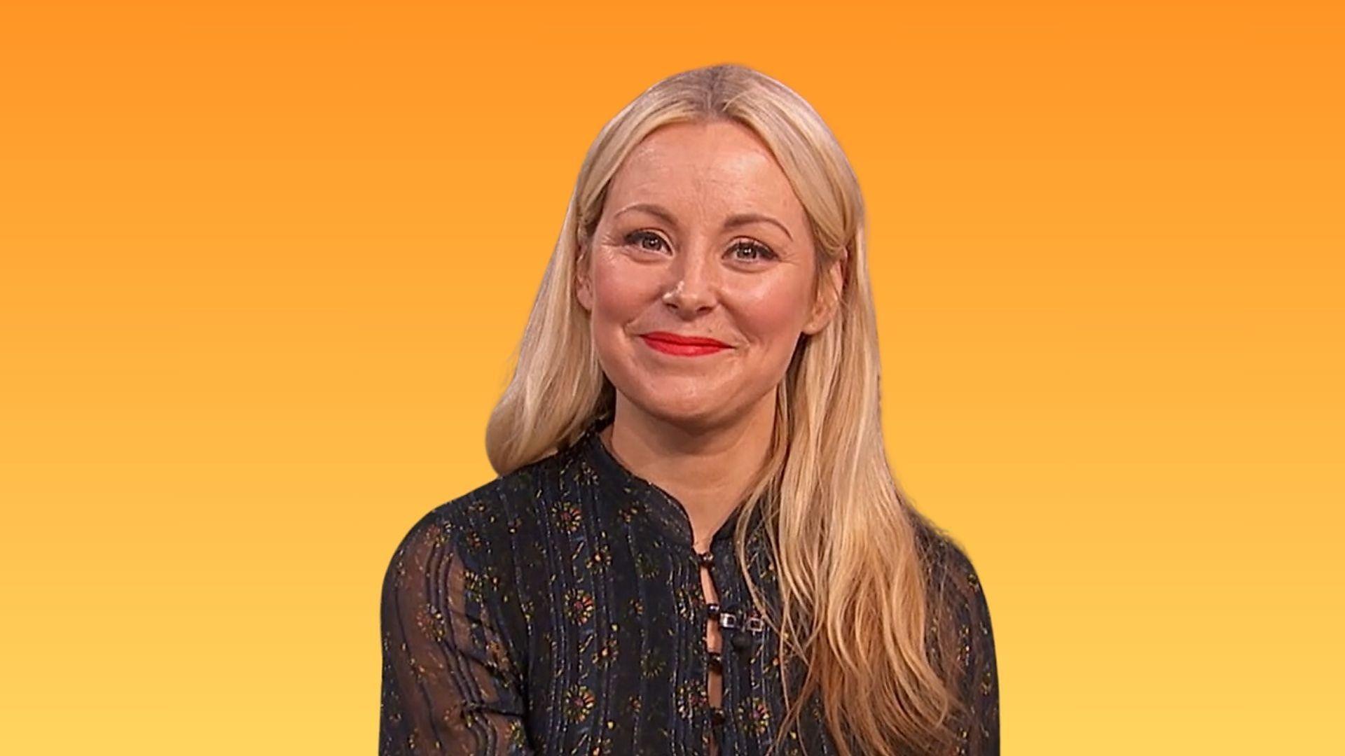 Close up image of Anna Haugh. She wears a black top and is before a yellow and orange gradient background. Her blonde hair is down and she smiles at the camera.