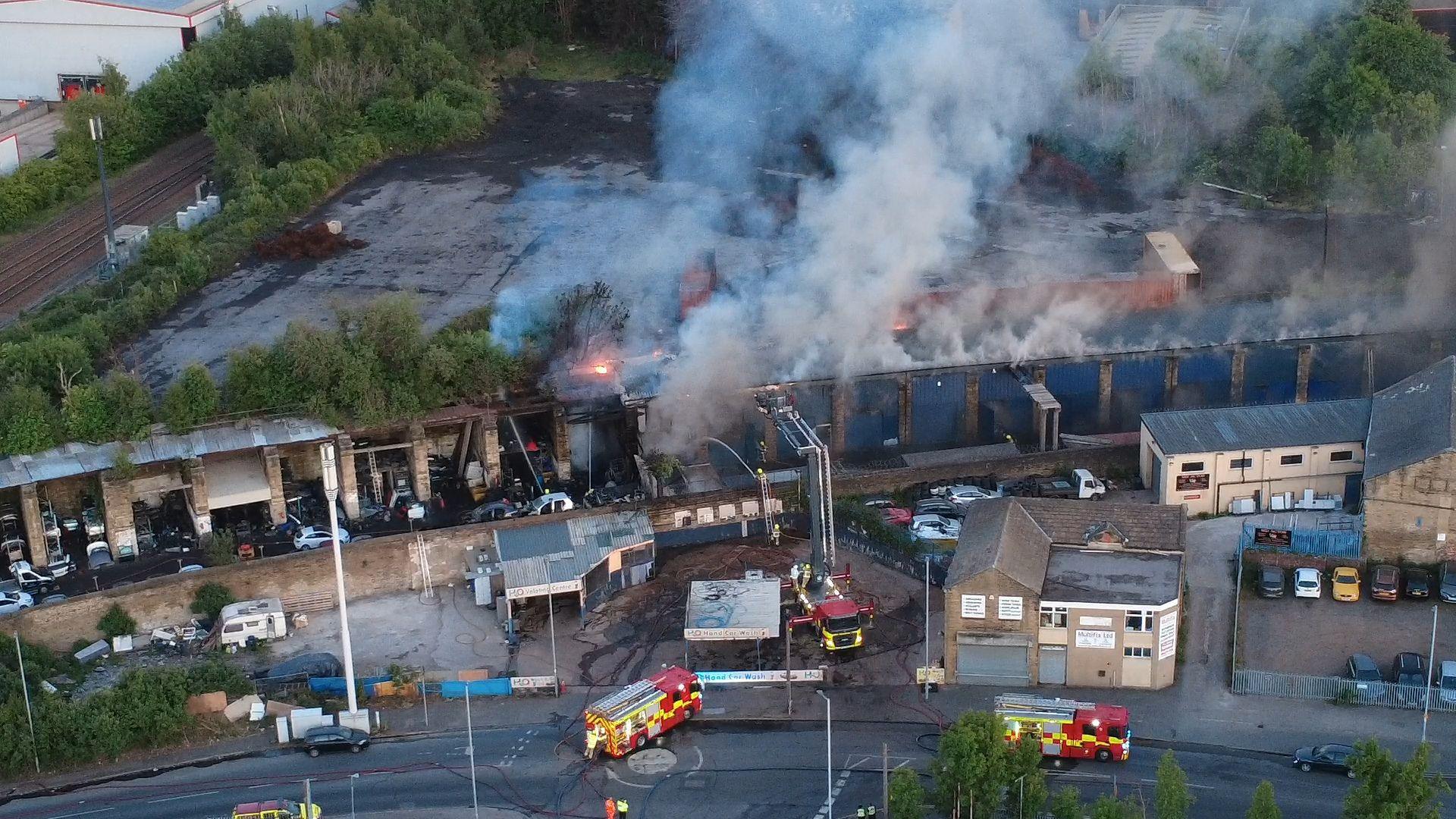 An aerial shot of a scrap yard on fire with several fire engines around it