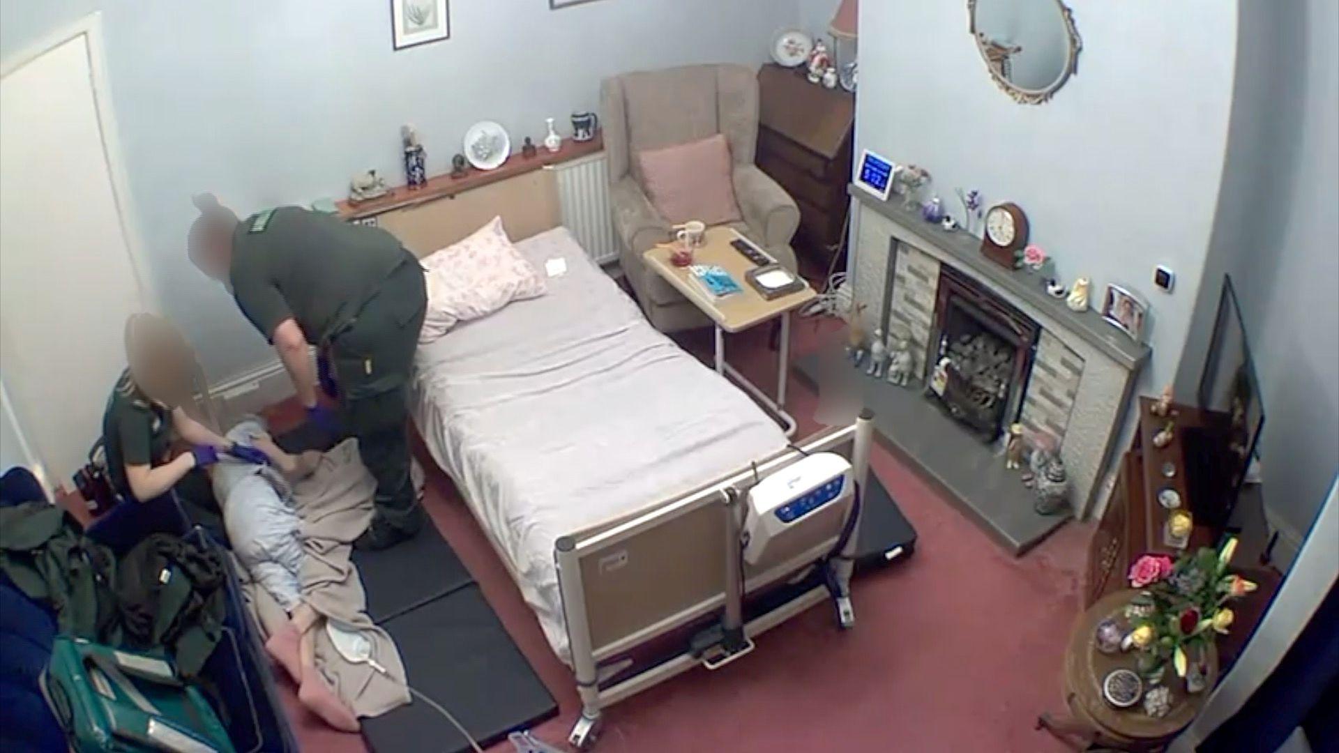 A CCTV image show an elderly woman lying on a living room floor next to a hospital bed.  She is being helped by paramedics.  The paramedics have their faces blurred out.  You can't see the face of the woman lying on the floor.
