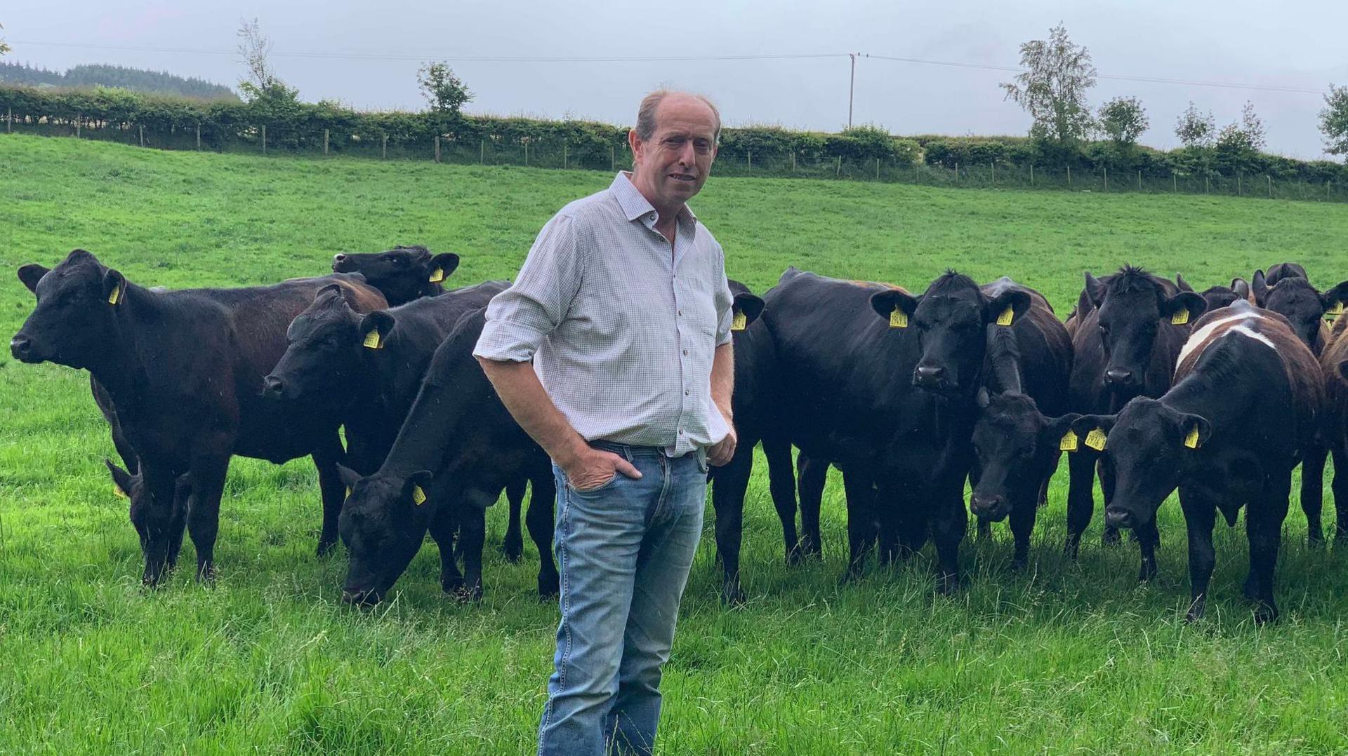 Farmer Ian Buchannan pictured in a light white-coloured  shirt and jeans while standing in front of a number of cows in a field. 