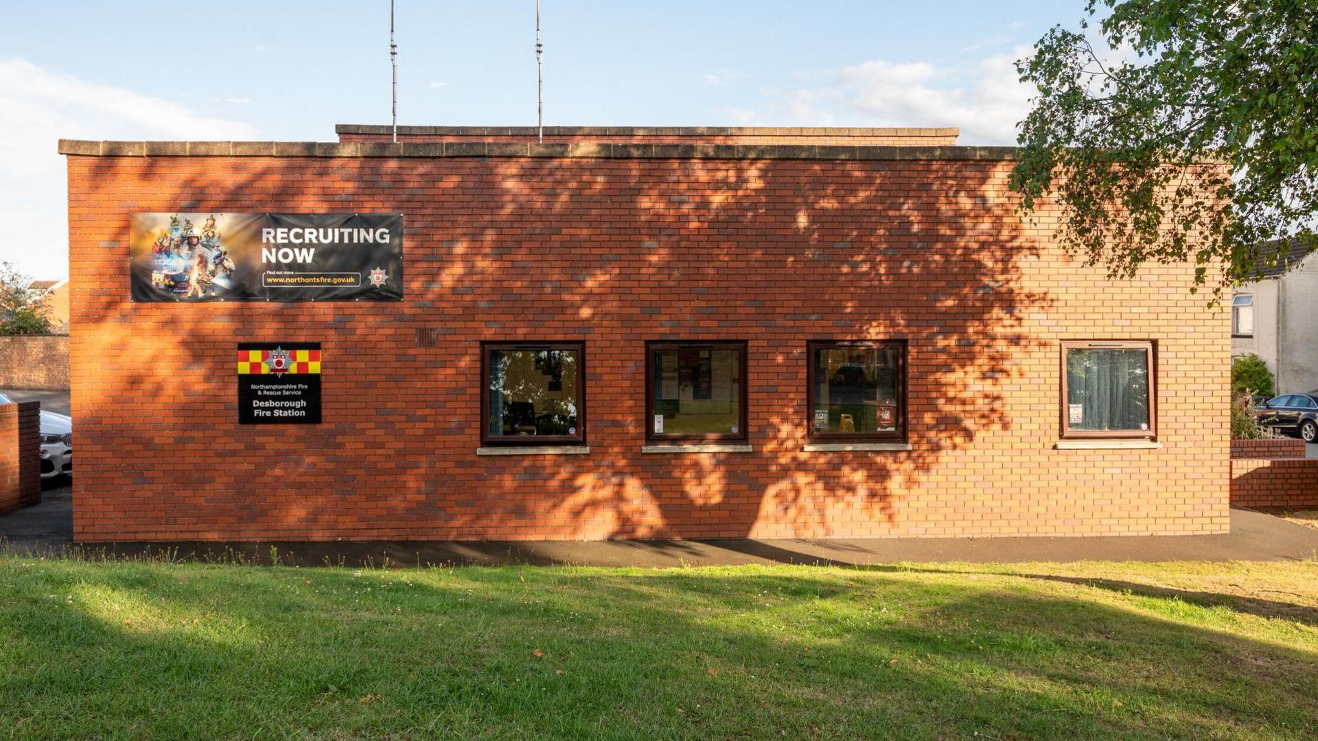 The outside of Desborough fire station, made of red brick with four windows, and signs on the building to the left. There is grass in front of the building and a tree to the right. 