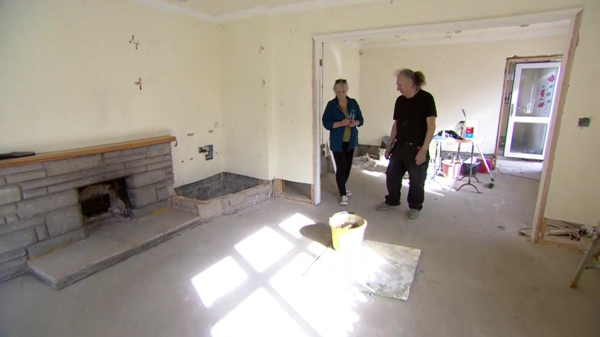 A woman and a man survey their flood-damaged bungalow. It has bare walls and floors and is undergoing redecoration work.