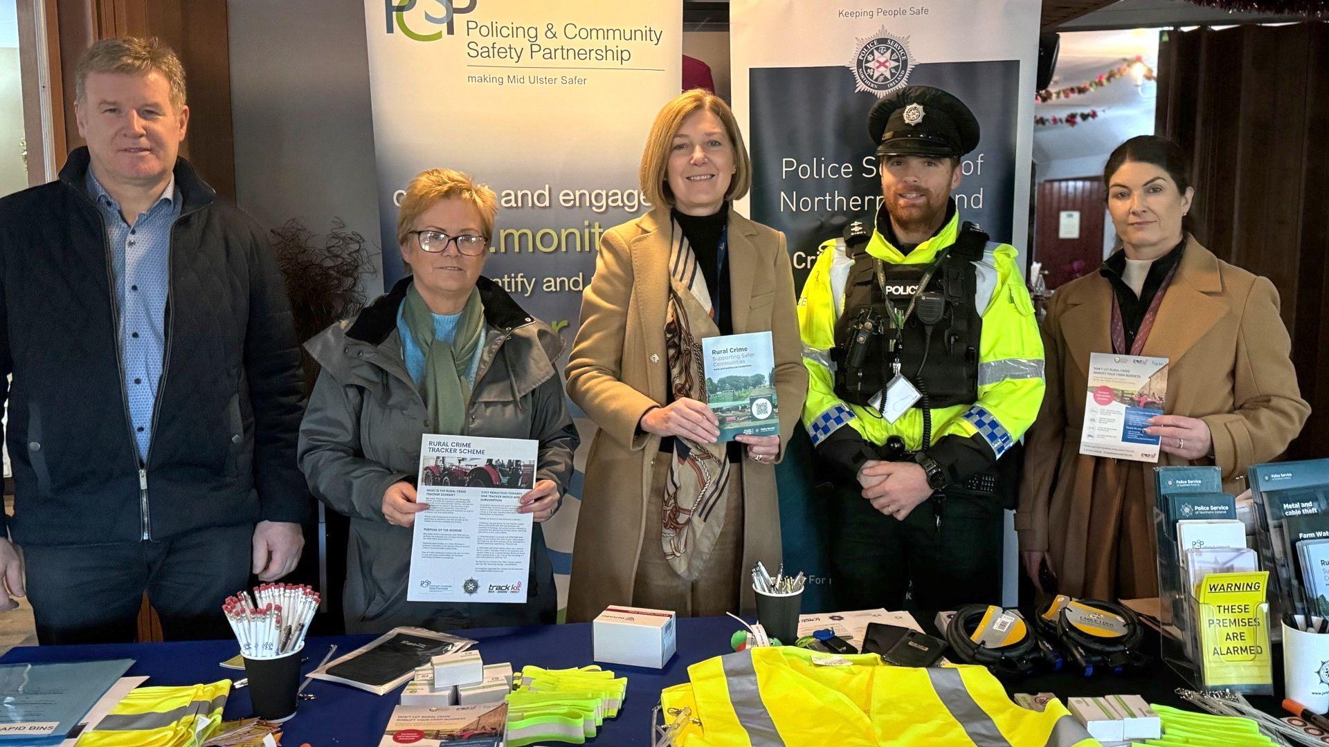 Five people - two men and three women - standing behind a table, some are holding leaflets. The table is full of stuff, including leaflets and hi-vis jackets. One of the men is a police officer who is in uniform.