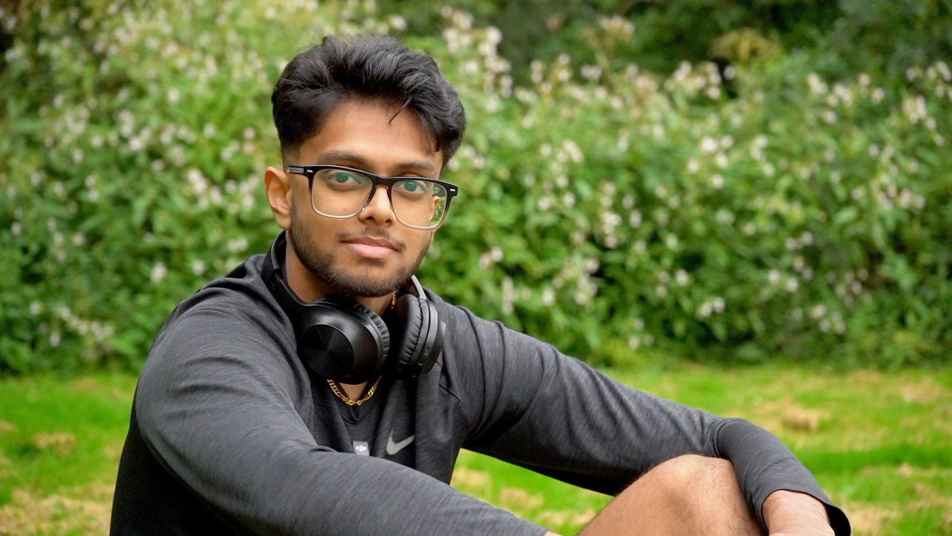 Mridul is sat in a field with his headphones around his neck looking into the camera.