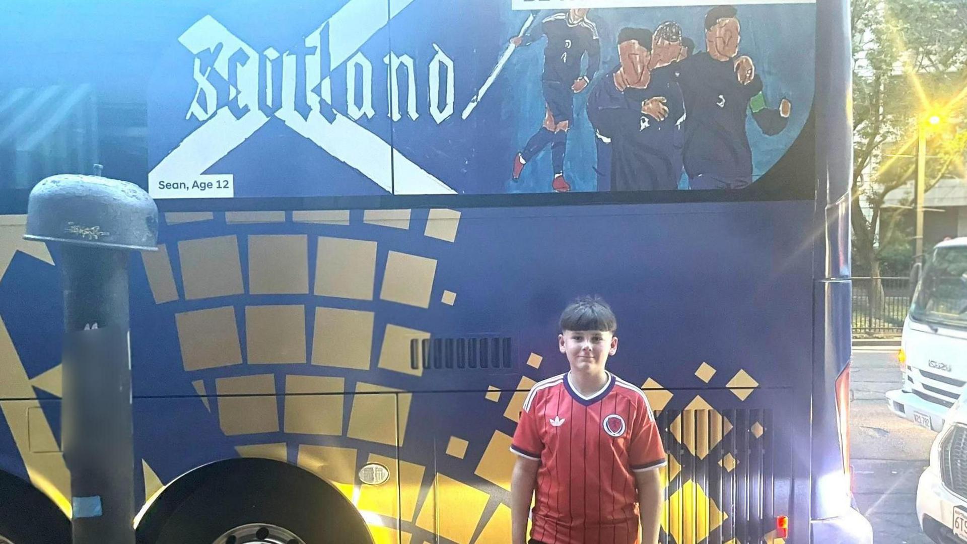 A boy wearing a red football top stands beside the Scotland World Cup team bus, which features his design.