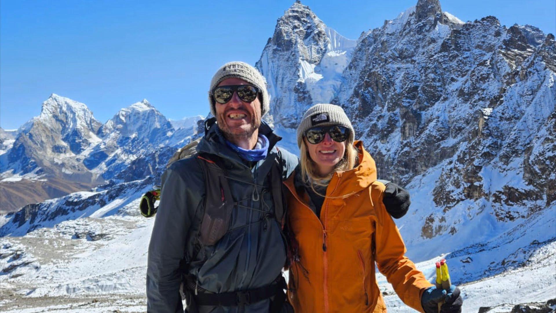 Rik and Liv have their arms around each other as they smile for a photo in front of a mountain with snow in the background. Liv is wearing a beanie hat, sunglasses and a orange coat and is holding hiking polies. Rik also has a beanie hat on and a dark blue coat.