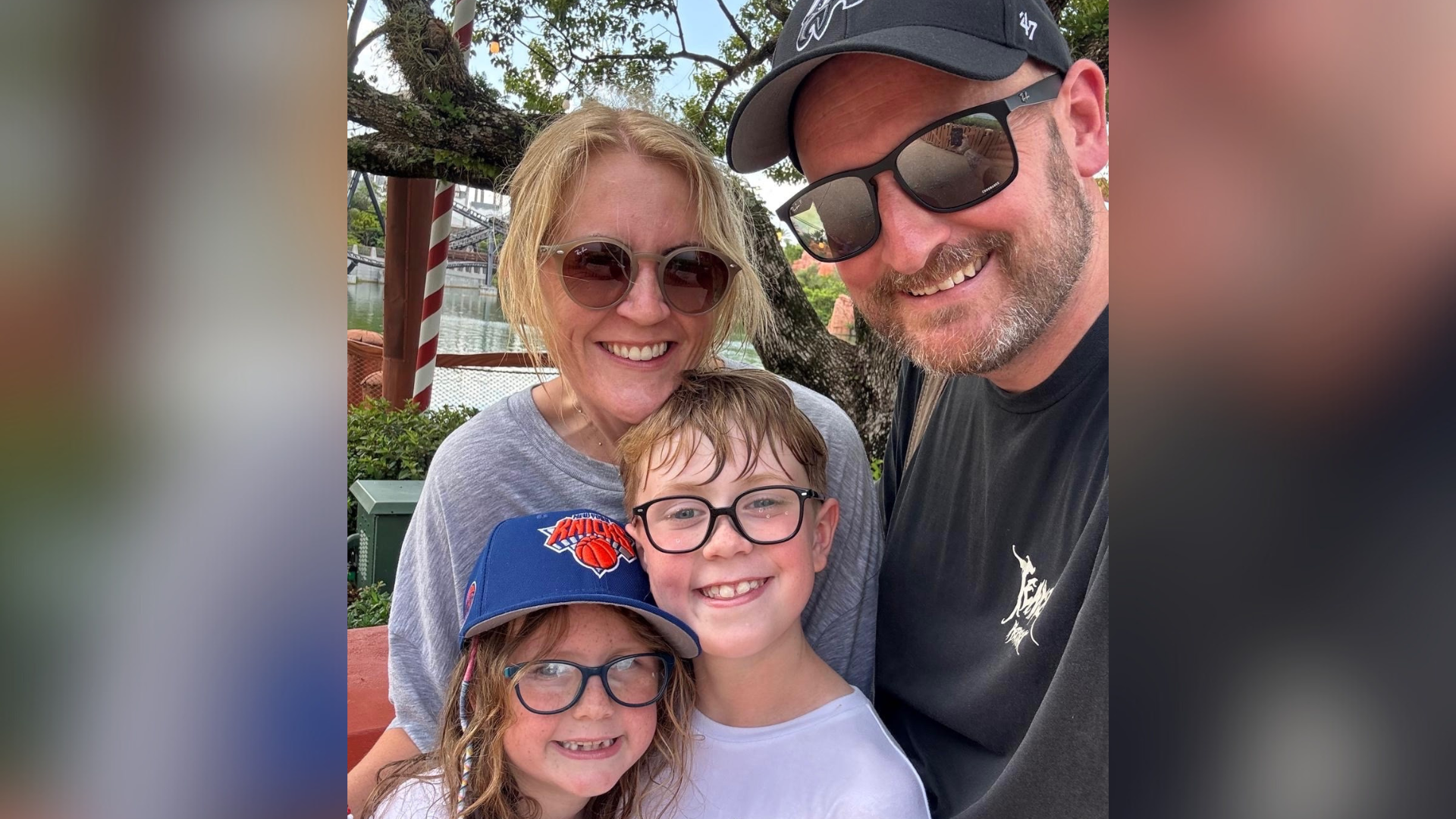 A cheerful family moment captured outdoors, featuring Mr Whitaker, his wife, and two children all wearing glasses. The group is smiling warmly, with one child is in a blue cap and the other appearing freshly out of water. Mrs Whitaker has long blonde hair and is wearing a grey shirt, while Mr Whitaker wears a black shirt and black baseball cap.