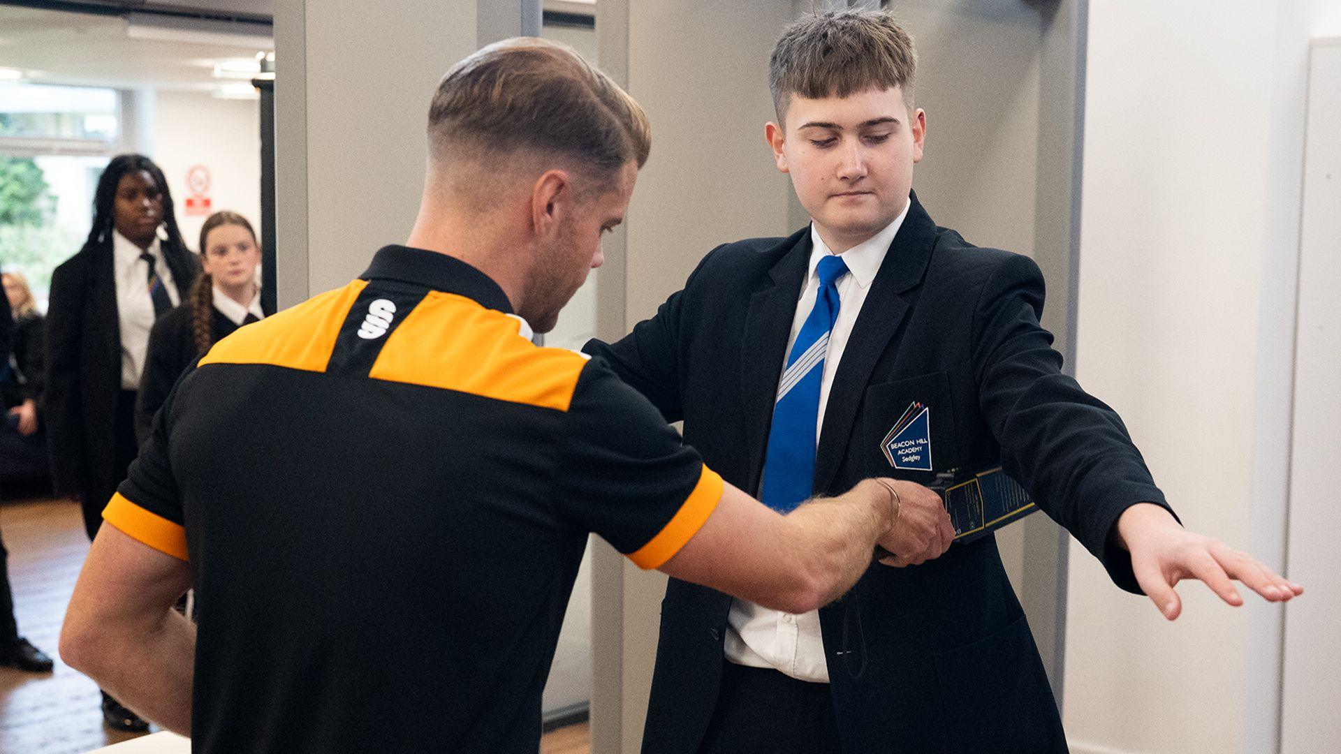 A school boy is scanned with an portable metal detector by a member of school staff. In the background children queue by a doorway. 