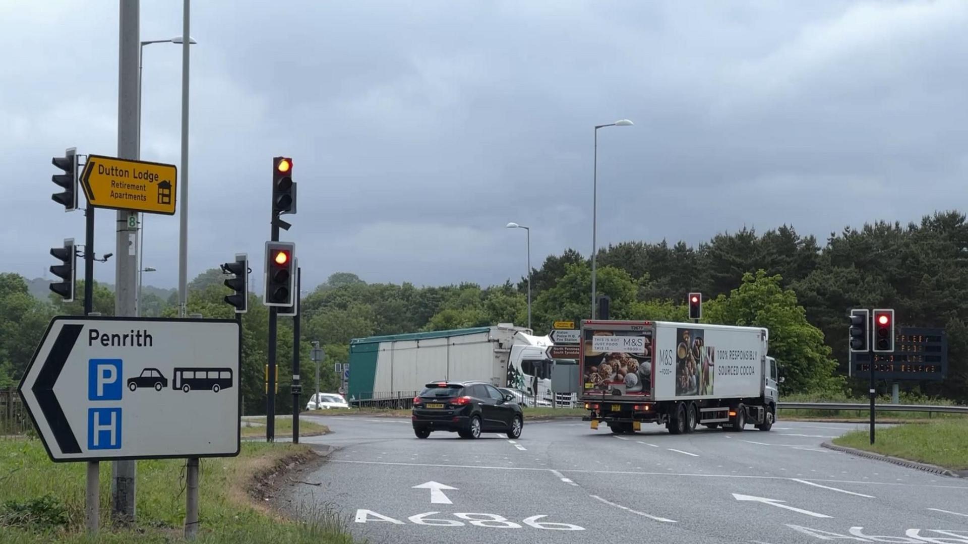 Vehicles are waiting at red traffic lights at Kemplay Bank roundabout, Penrith. There is an exit sign for Penrith at the left of the picture. A car and a lorry are waiting to exit the roundabout, heading east, while a lorry and car are waiting to enter from the A686.