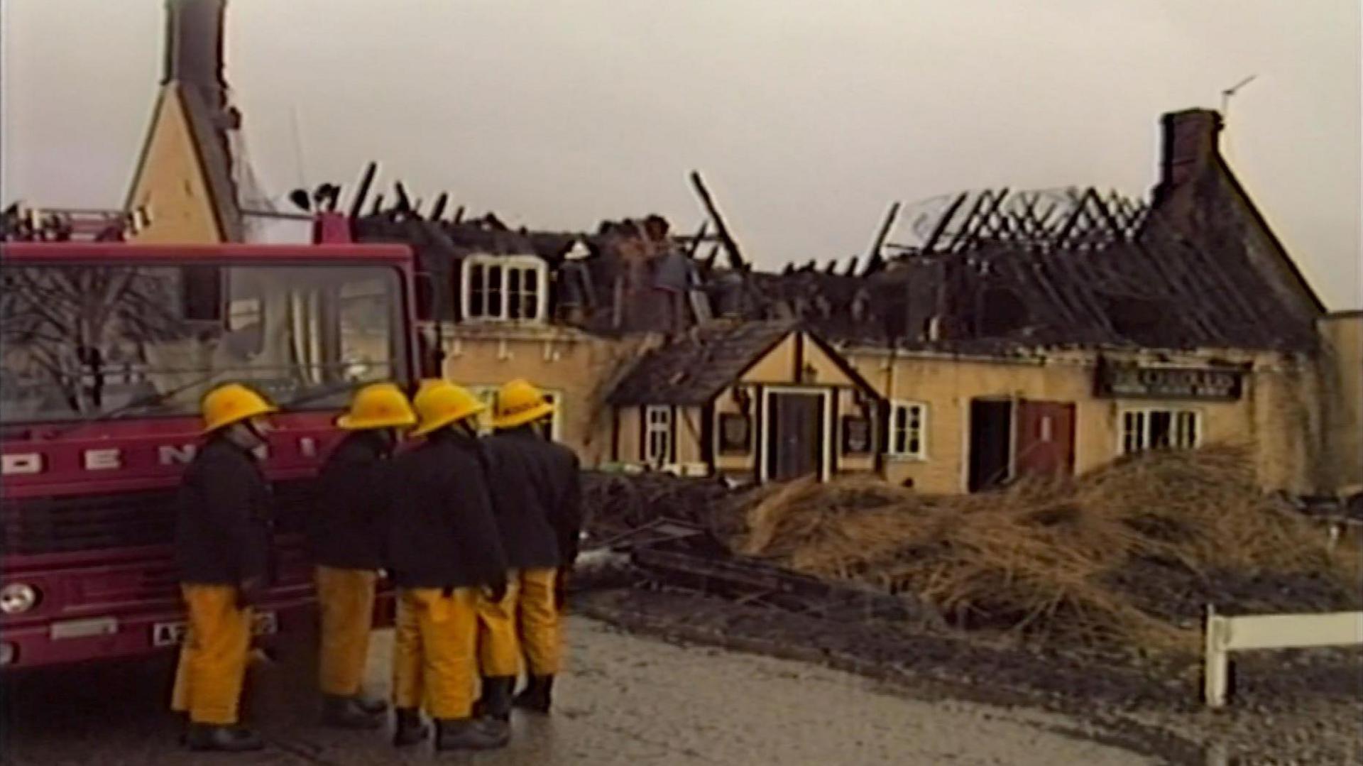 An old news photo shows four Norfolk firefighters standing beside a large red fire truck on a grey, overcast day. In the background, a large building has lost its roof and appears badly damaged by fire.