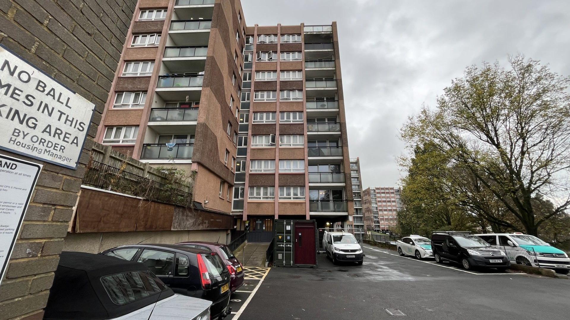 The outside of Kingfisher Court. There is a car park with a number of cars and taxis parked in front of a 12-storey block of flats. 