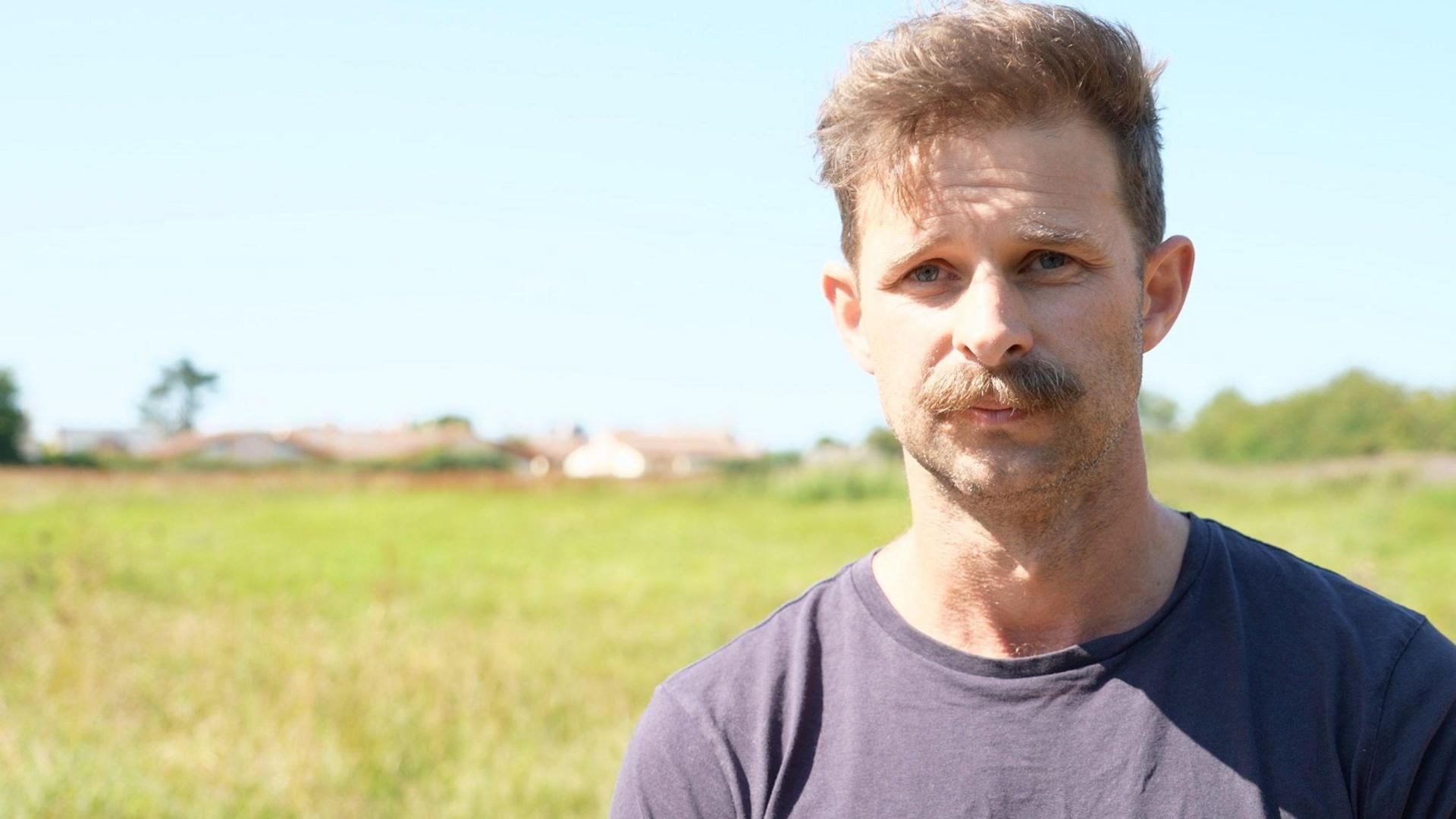 A man with short, light brown hair and a moustache is standing outdoors in a grassy field. The background shows a clear blue sky and some distant buildings and trees.