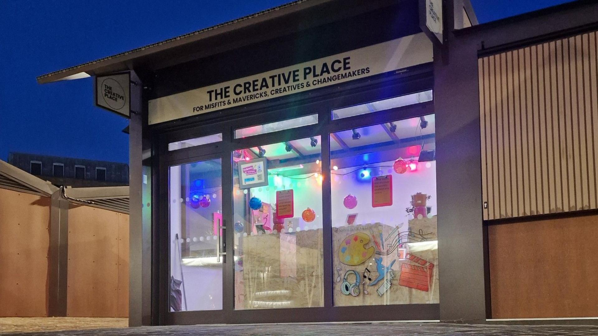 The exterior of a wooden market stall on which is the legend "The Creative Place" in black capitals. There are three windows below on which are posters about events.