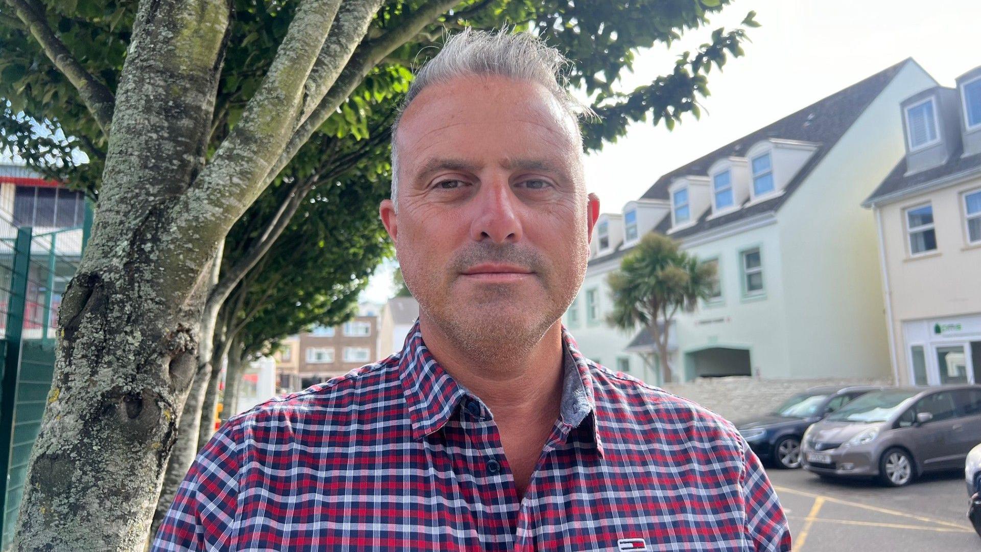 Carl Walker is wearing a red chequered shirt, he has grey hair and is stood under a tree