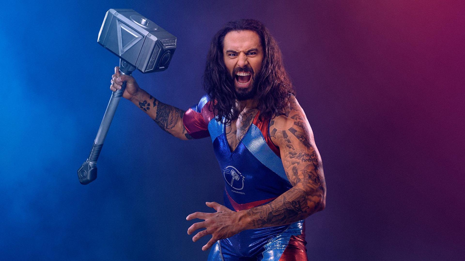 A main with long hair, wearing a bright red and blue shiny outfit, holds a hammer and shouts into the camera