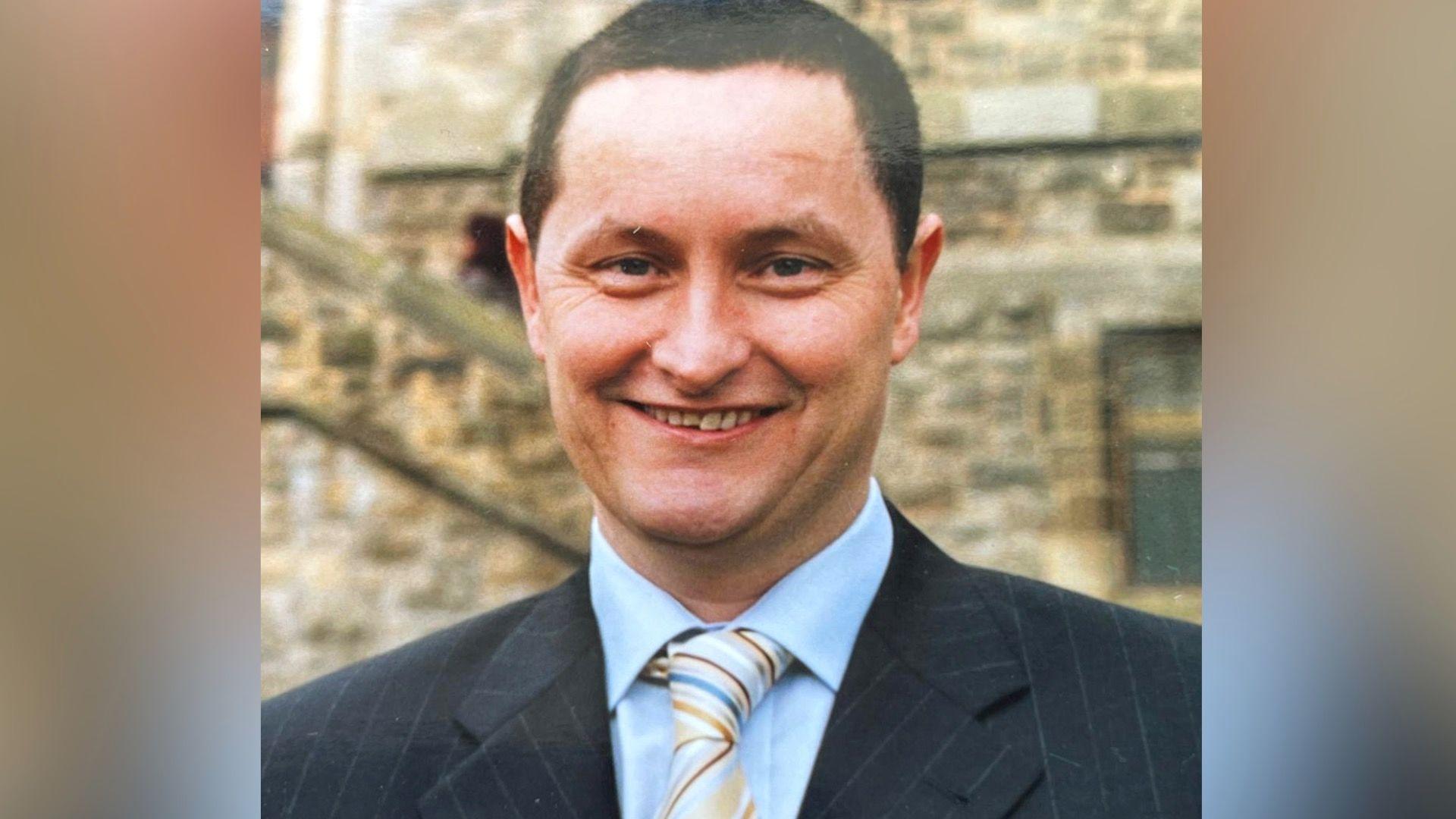 A younger John Kelly wearing a suit with a blue shirt and multi-coloured stripey tie. He has dark hair and is smiling at the camera