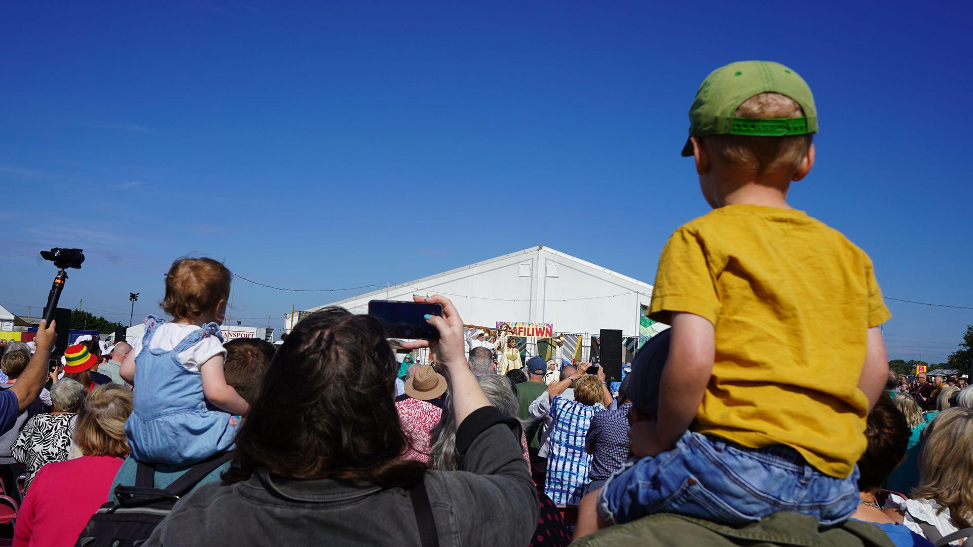 Plentyn ifanc ar ysgwyddau oedolyn yn gwylio seremoni