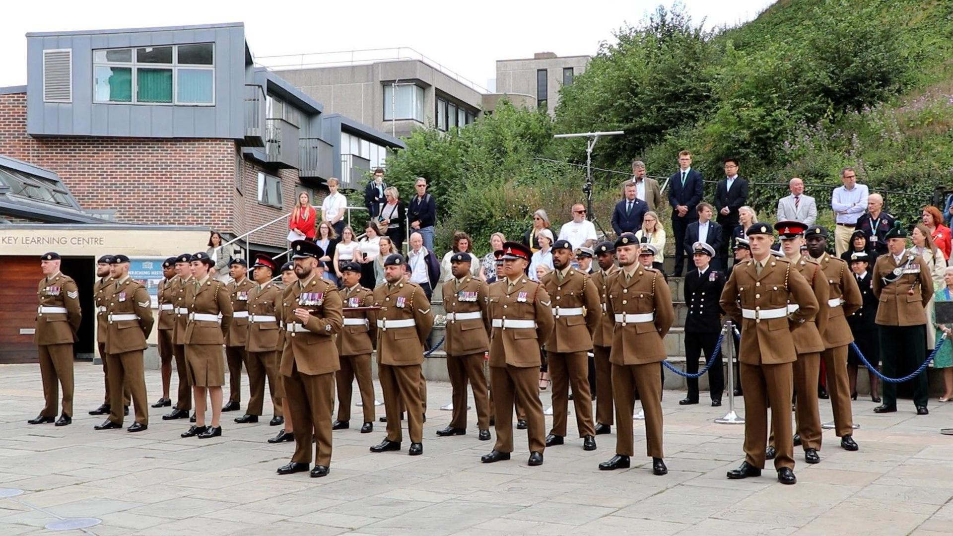 Military personnel in official clothing stand in front of a large grass mound.