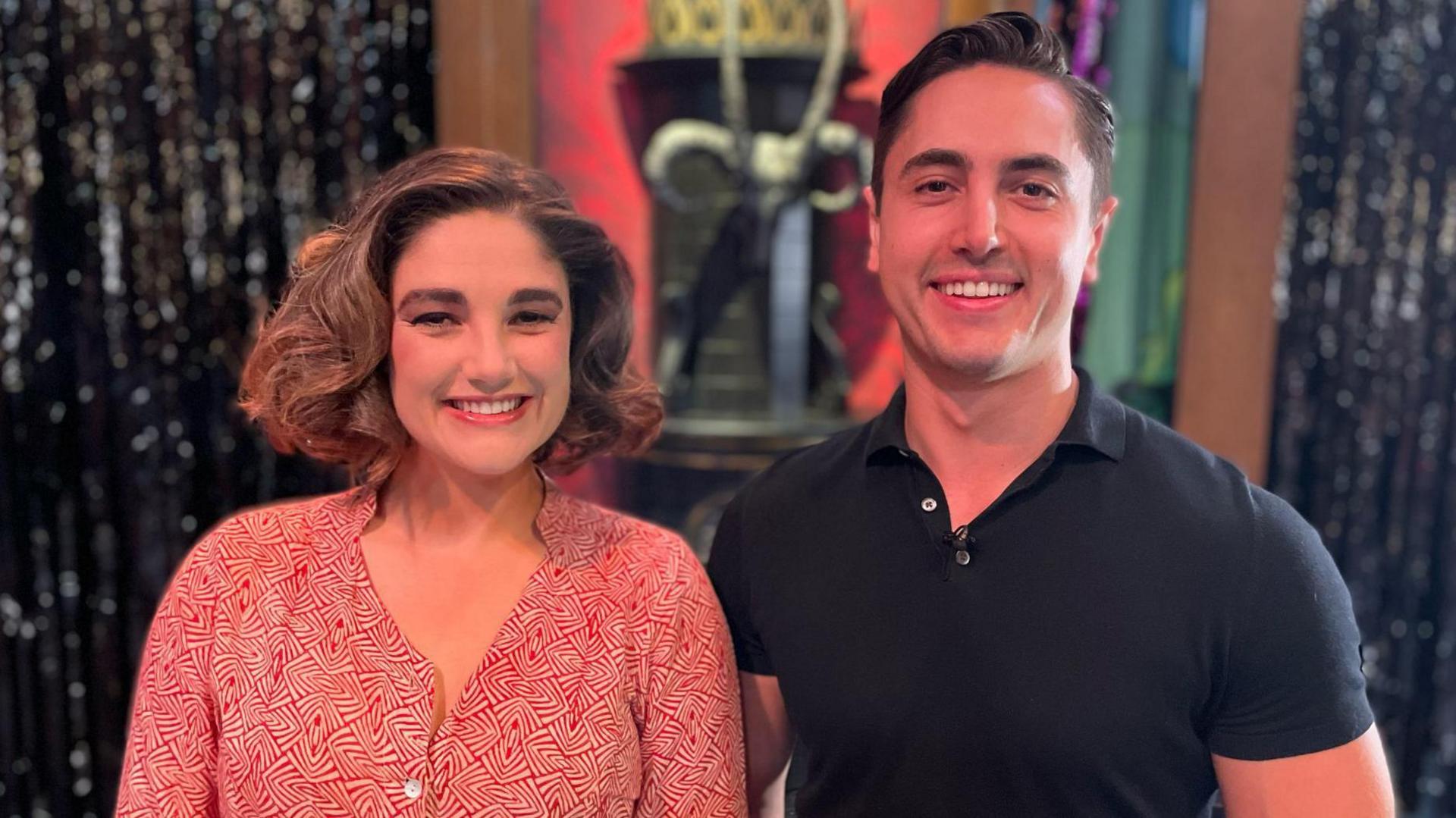 Natasha is in the red patterned dress and has curled her hair. Connor Carson is standing beside her in a tight black t-shirt. There are standing on the stage and some of the set is visible behind them
