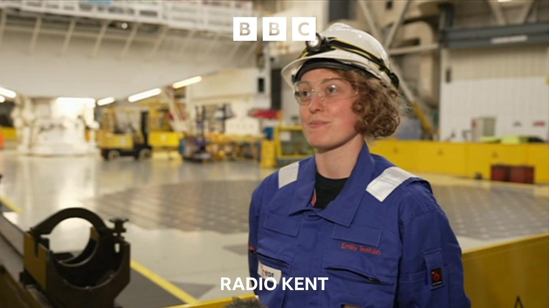 A young woman wearing a blue boiler suit and white hard hat stands in a large open room. technical equipment and yellow barriers can be seen in the background.