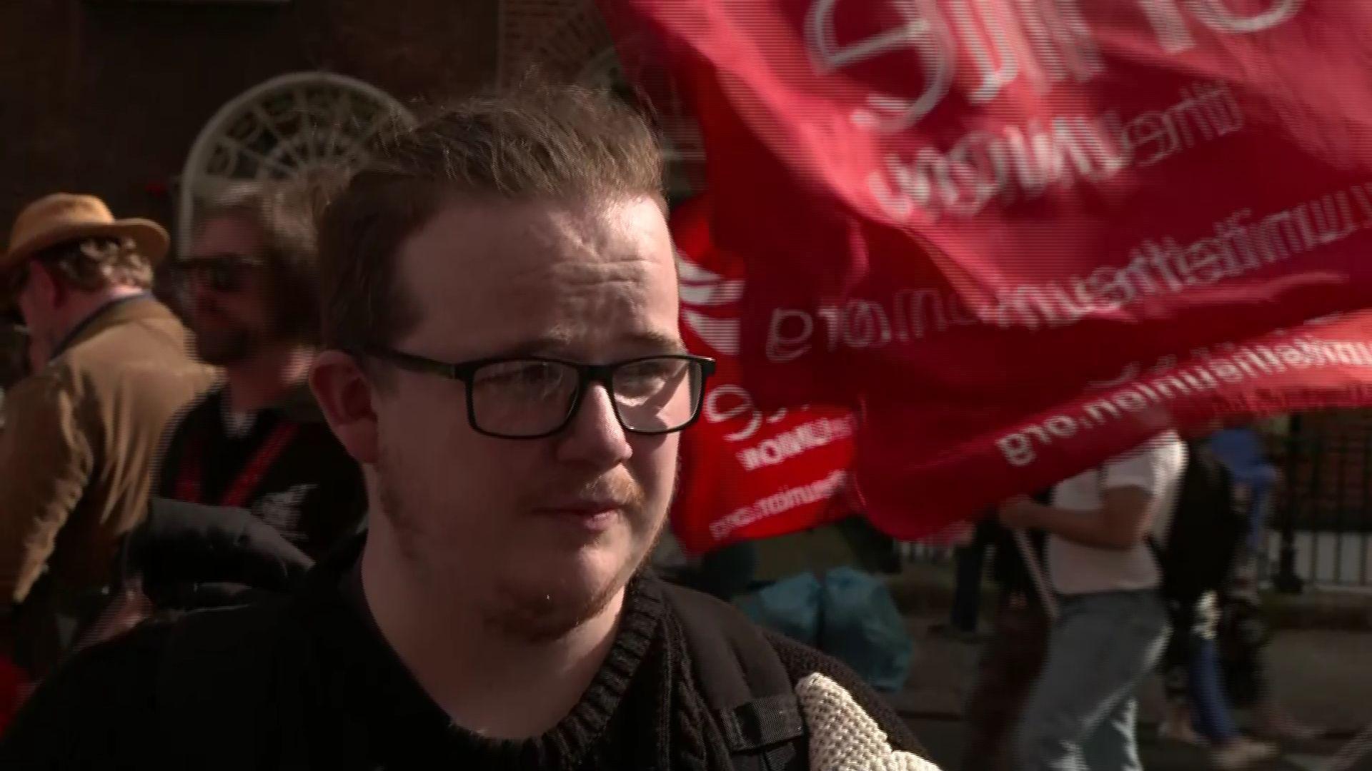 A young man with brown hair pulled back off his face. He is wearing black framed glasses, a dark jumper and has some facial hair.