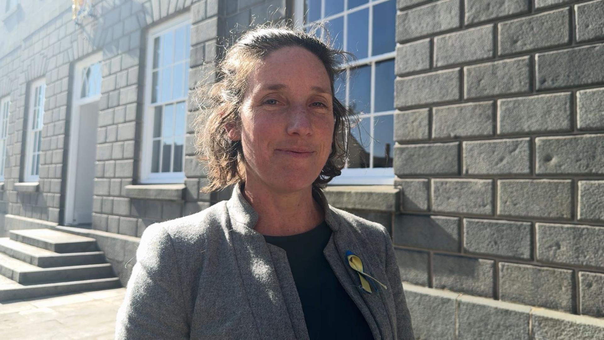 A woman wearing a grey jacket with a Ukraine flag pin. Her brunette bob is half pinned back. She is stood in front of the States of Guernsey building. 