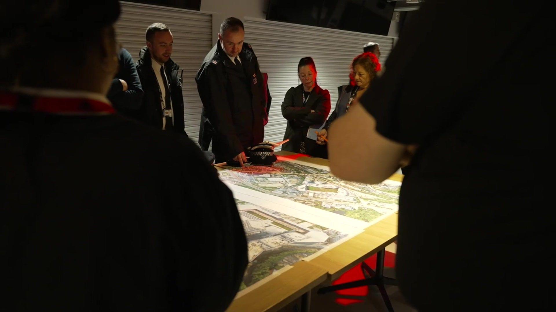 A group of officials, including police, stadium staff., gathers around a large map on a table