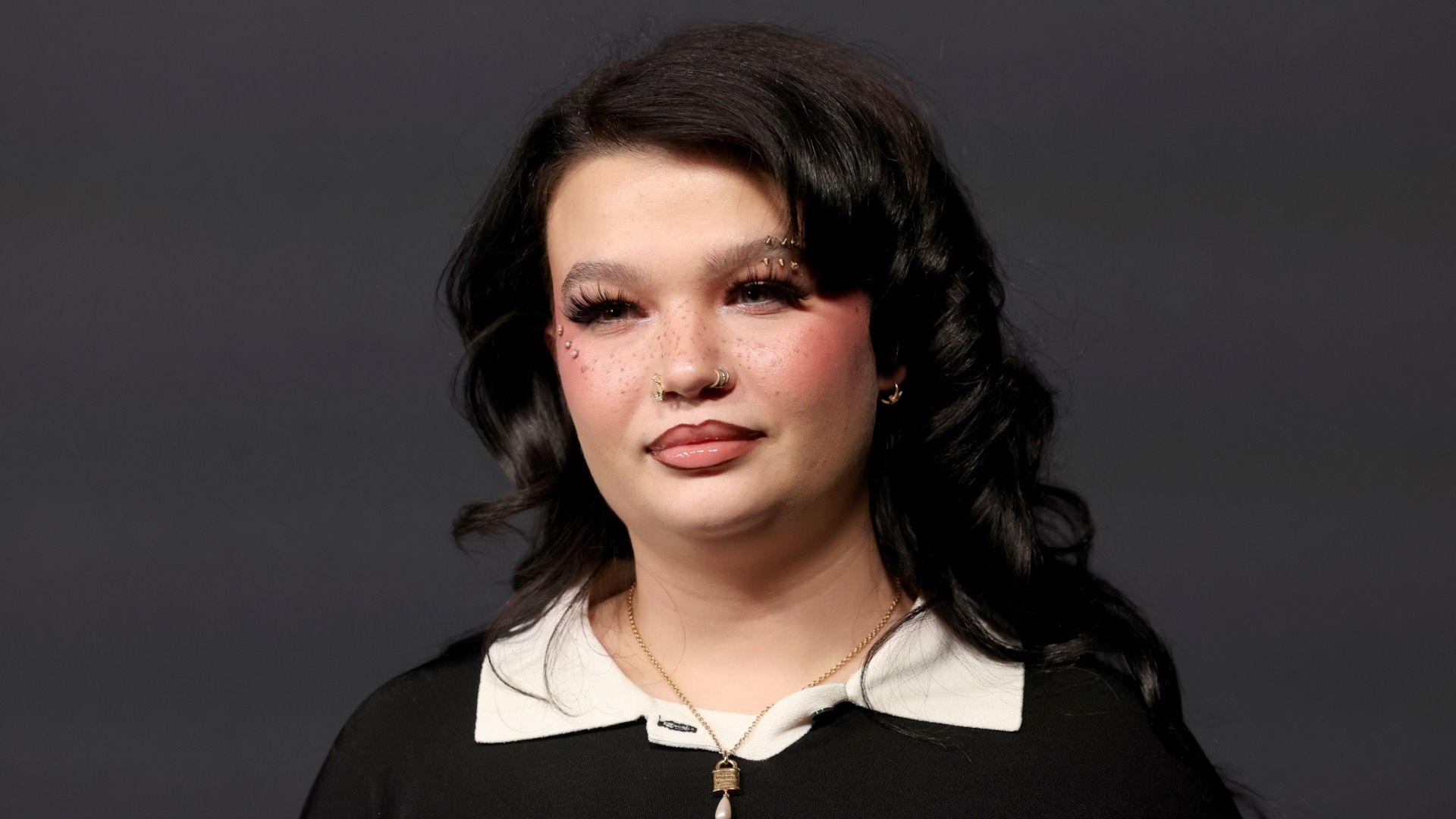 Lola Young smiles as she stands in front of a grey background, wearing a black top with a cream collar and lots of sparkly jewellery.