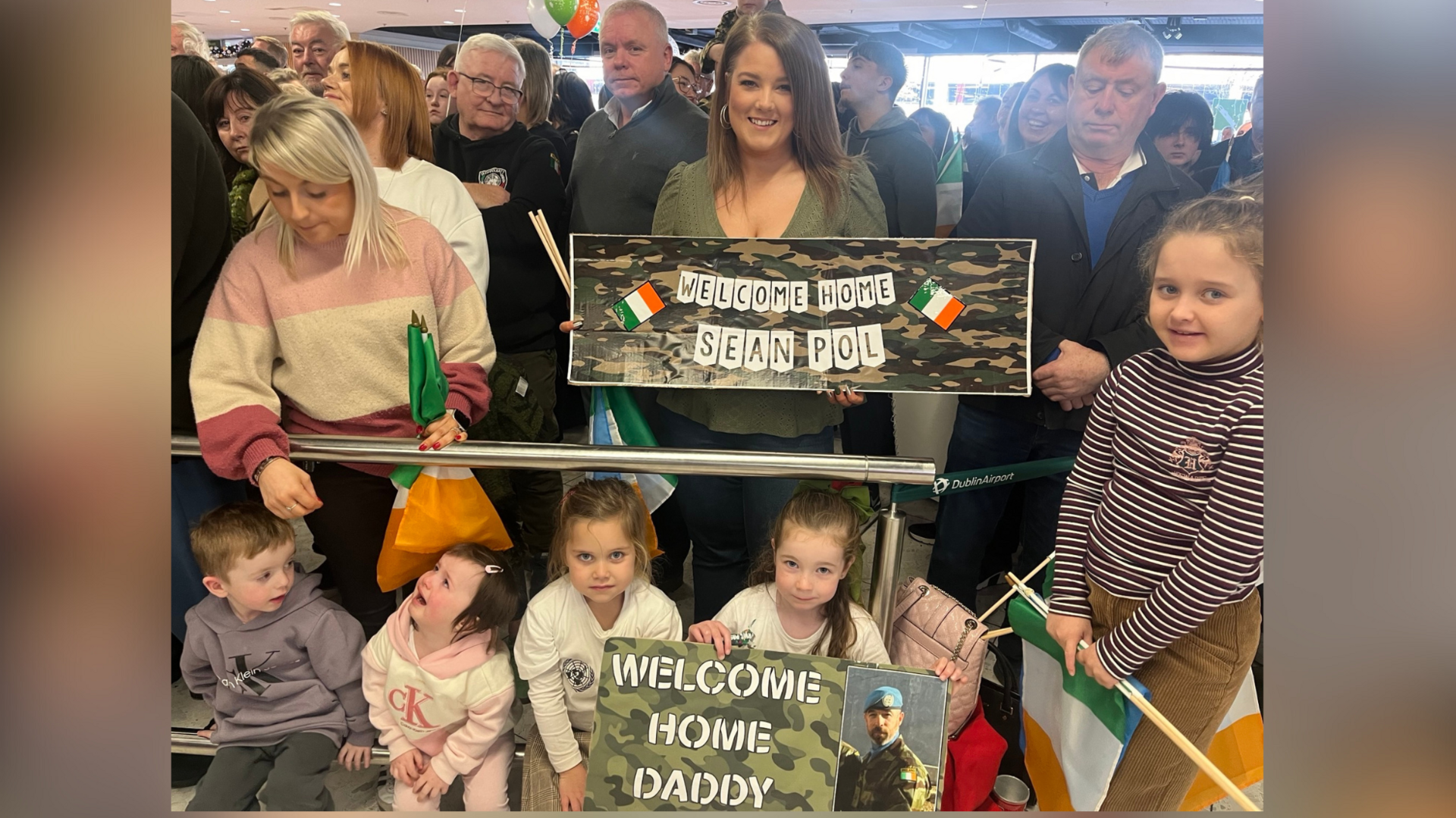 A woman stands smiling in a green top with a banner that says welcome home Sean Pol. There are Irish flags on the sign. Below her are children in white tops that hold a sign which says welcome home daddy.