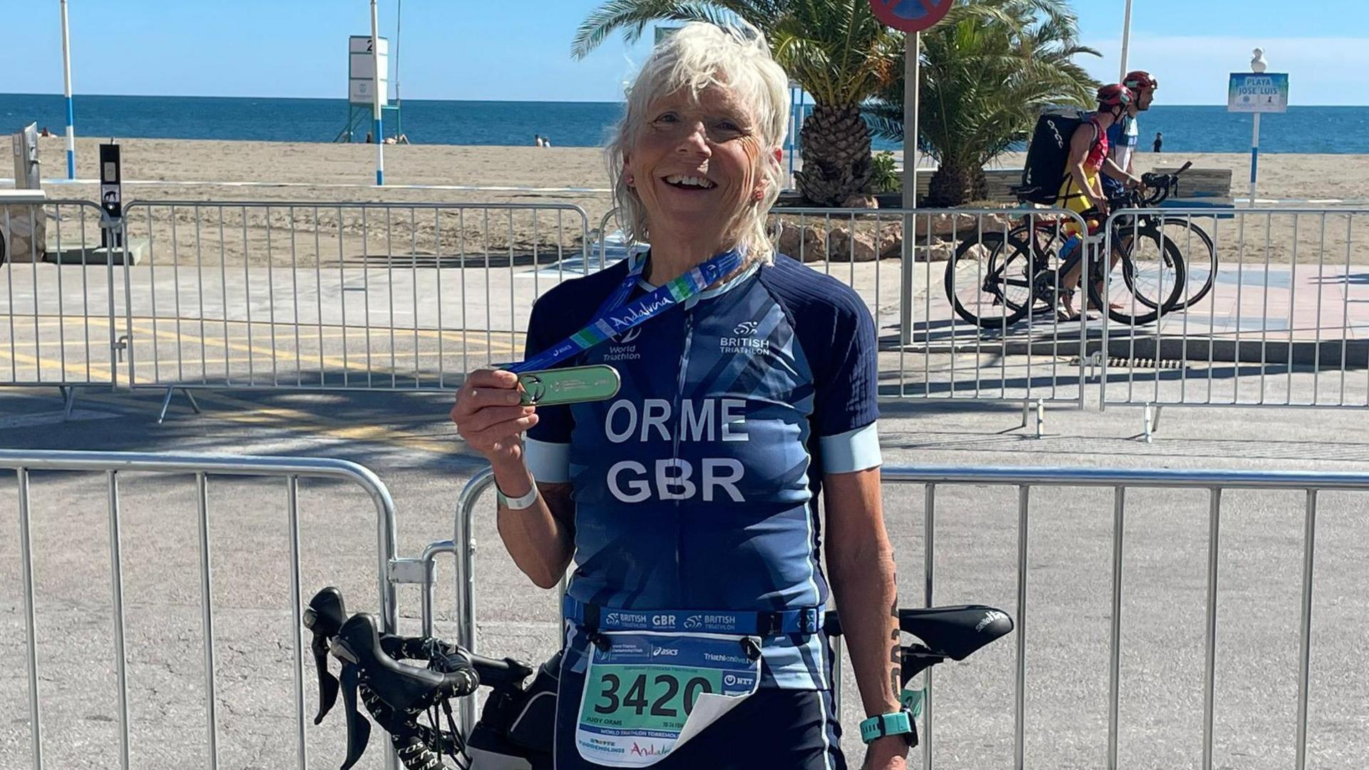 Judy holding her medal which is draped around her neck. Her bike is leaning on a metal fence. She is centre and smiling into the camera. A beach and the sea is in the background.