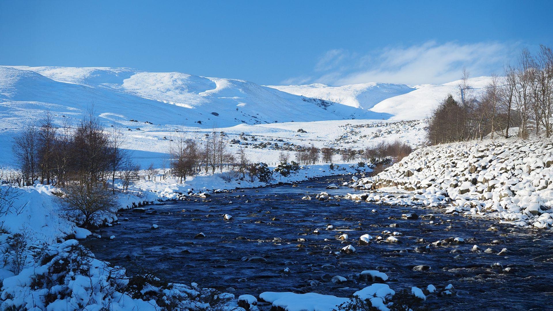 A wide shallow river with rocks protruding from the surface curves around a small hill. All of the ground is covered in snow with the large hills in the distance similarly encased in white. There are a number of brown, stripped bare trees lining the riverside. The sun is shining and the sky is a light blue colour.
