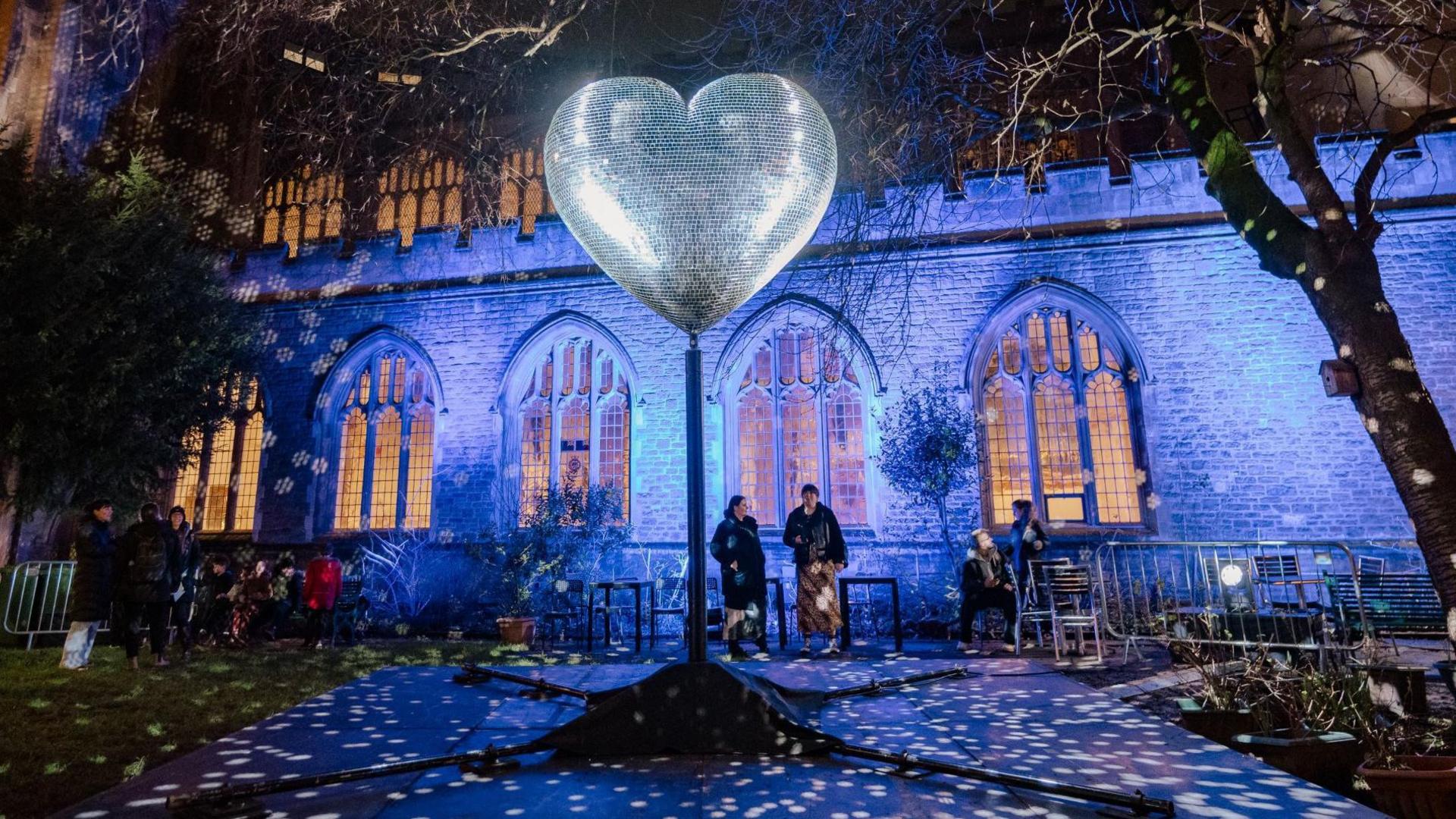 A large silver heart, designed to represent a disco ball, glows in the dark in a churchyard in central Bristol. In the background the main church building is illuminated in blue lights as part of the 2025 Bristol Light Festival and a small group of people are looking at the heart