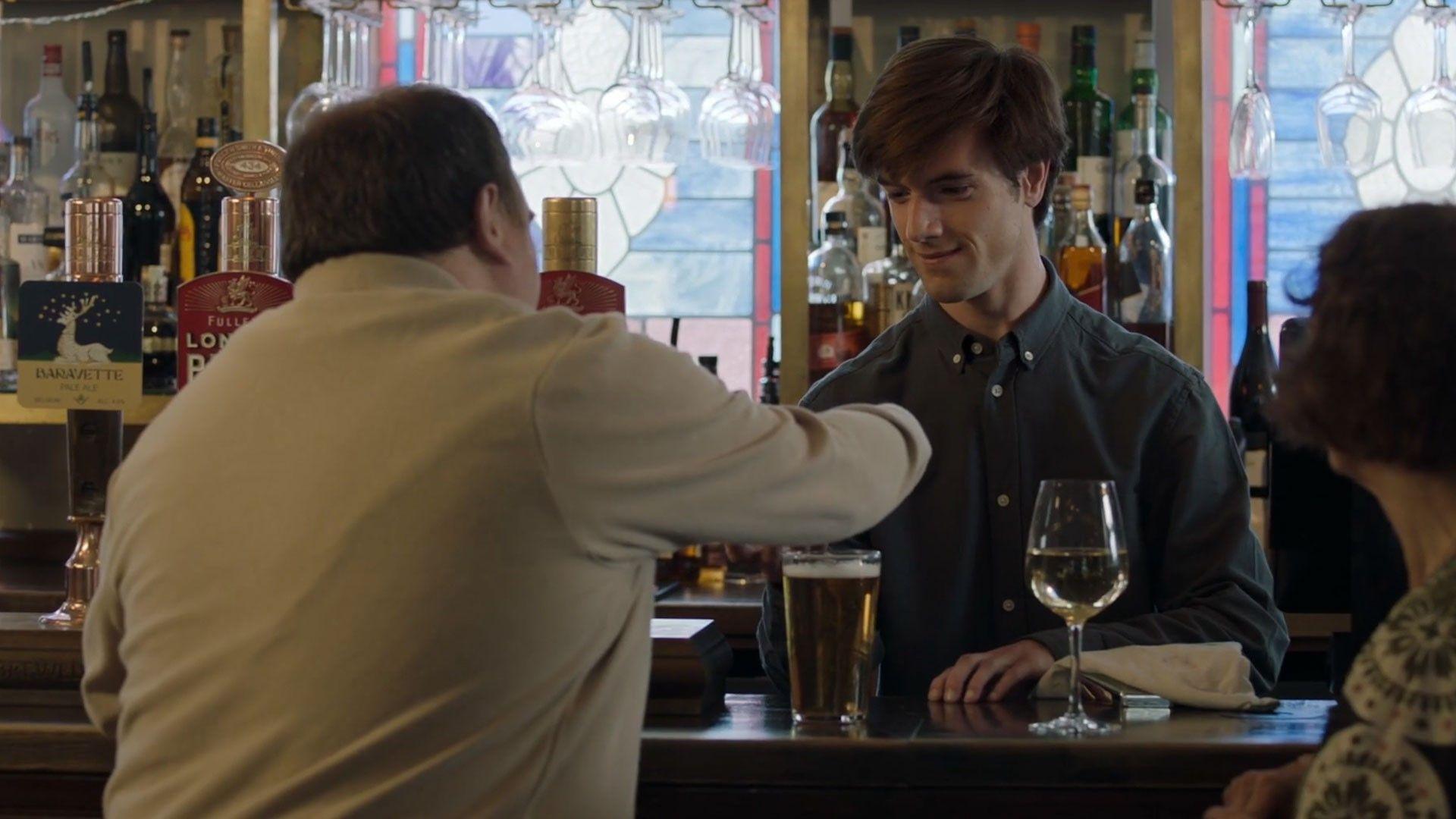 Jude acts as the barman character in The Chelsea Detective. He stands behind the bar of a pub taking payment from a man whose back we can see. Jude is smiling. There is a pint of beer and a glass of white wine on the bar.