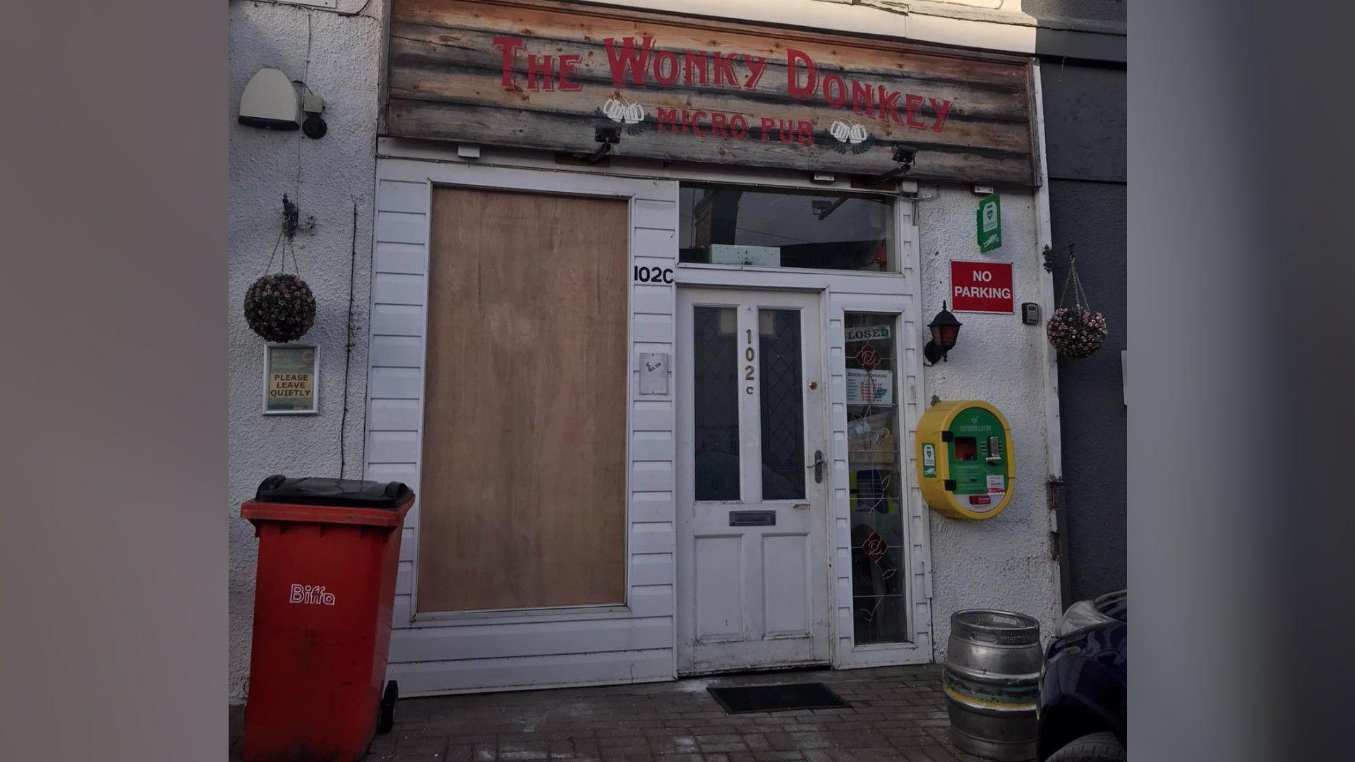 Frontage of the pub building- with a white door and a boarded up window- a red bin in front and a green and yellow defibrillator on the side of the building 