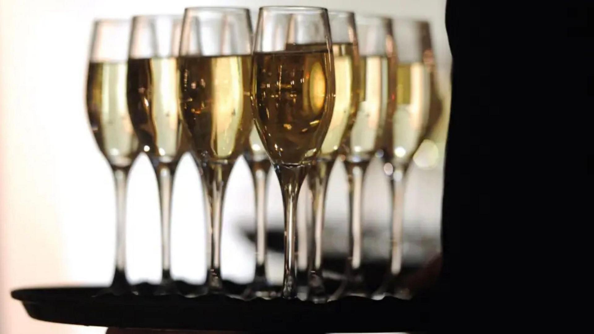 A tray of full champagne flutes filled with golden liquid. The stay is and hand holding is in silhouette. 