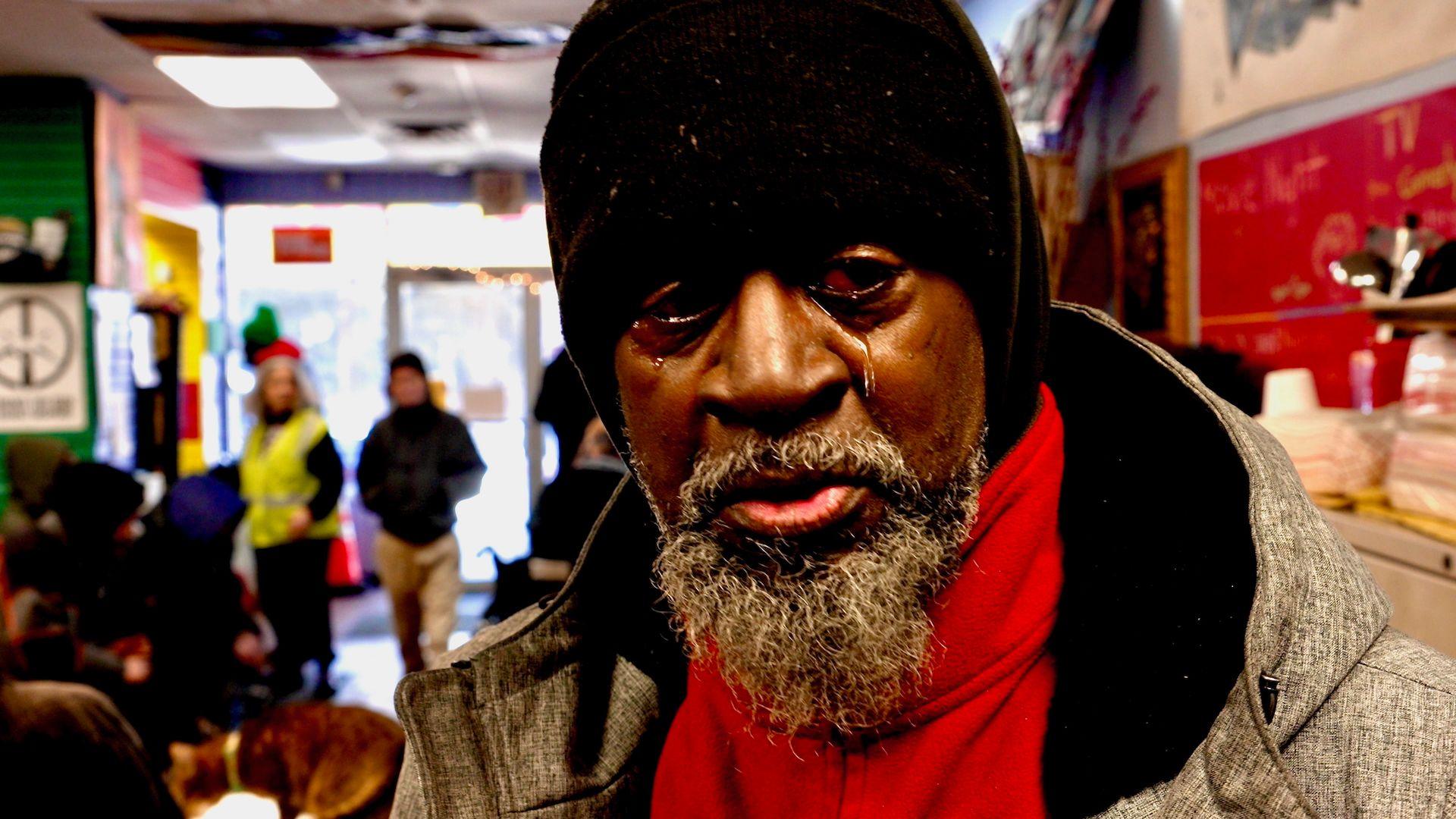 John White, a black man with a greying beard, who wears a dark woollen hat, a large grey coat and a red fleece underneath, photographed close-up at the drop-in centre with other attendees or staff visible out-of-focus behind him. A tear is rolling down his cheek.