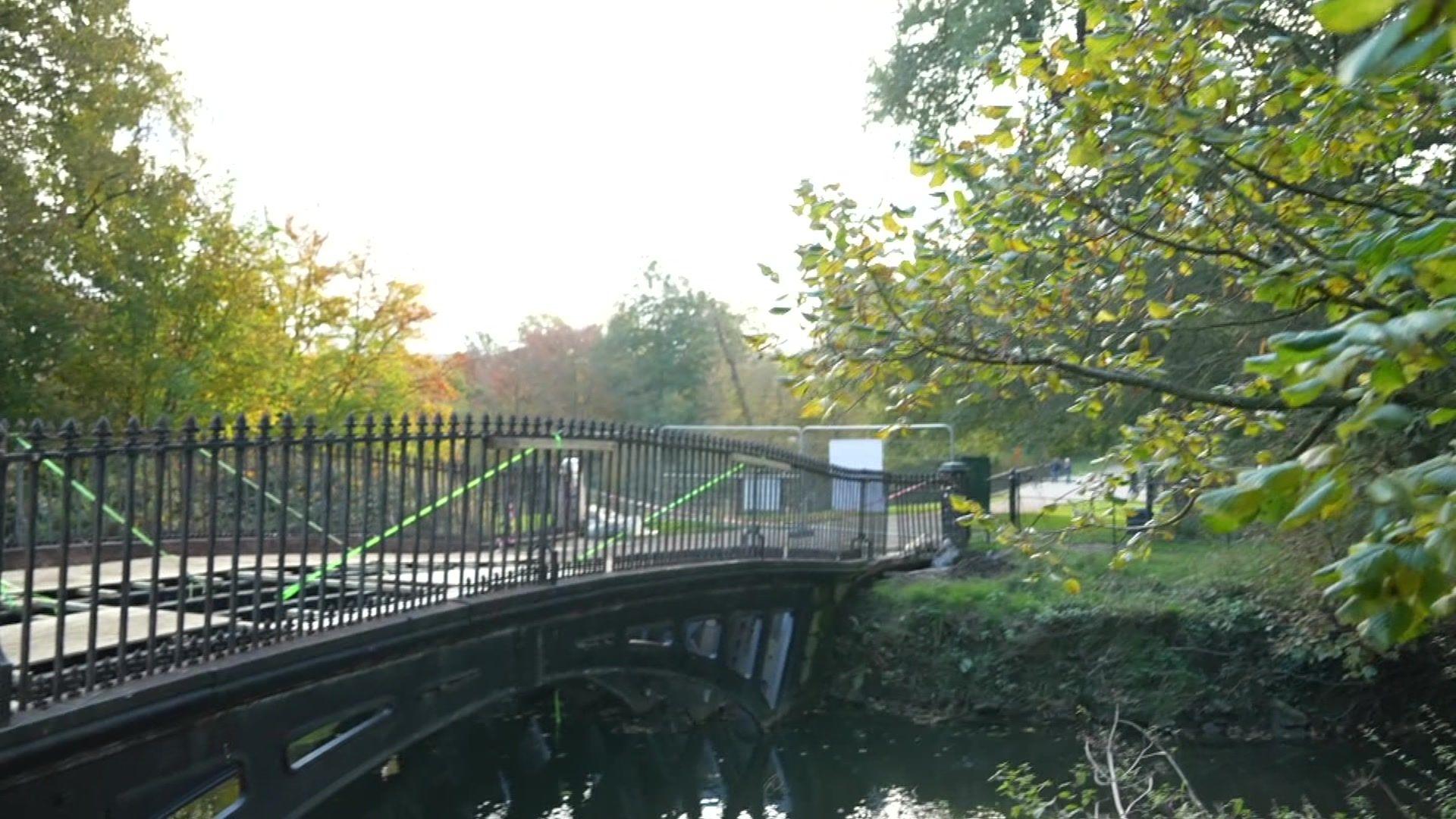 A cast iron bridge runs over a small stream, with trees flanking it on either side. Streams of green tape are stretched between the railings and the bridge floor, indicating construction work is happening.