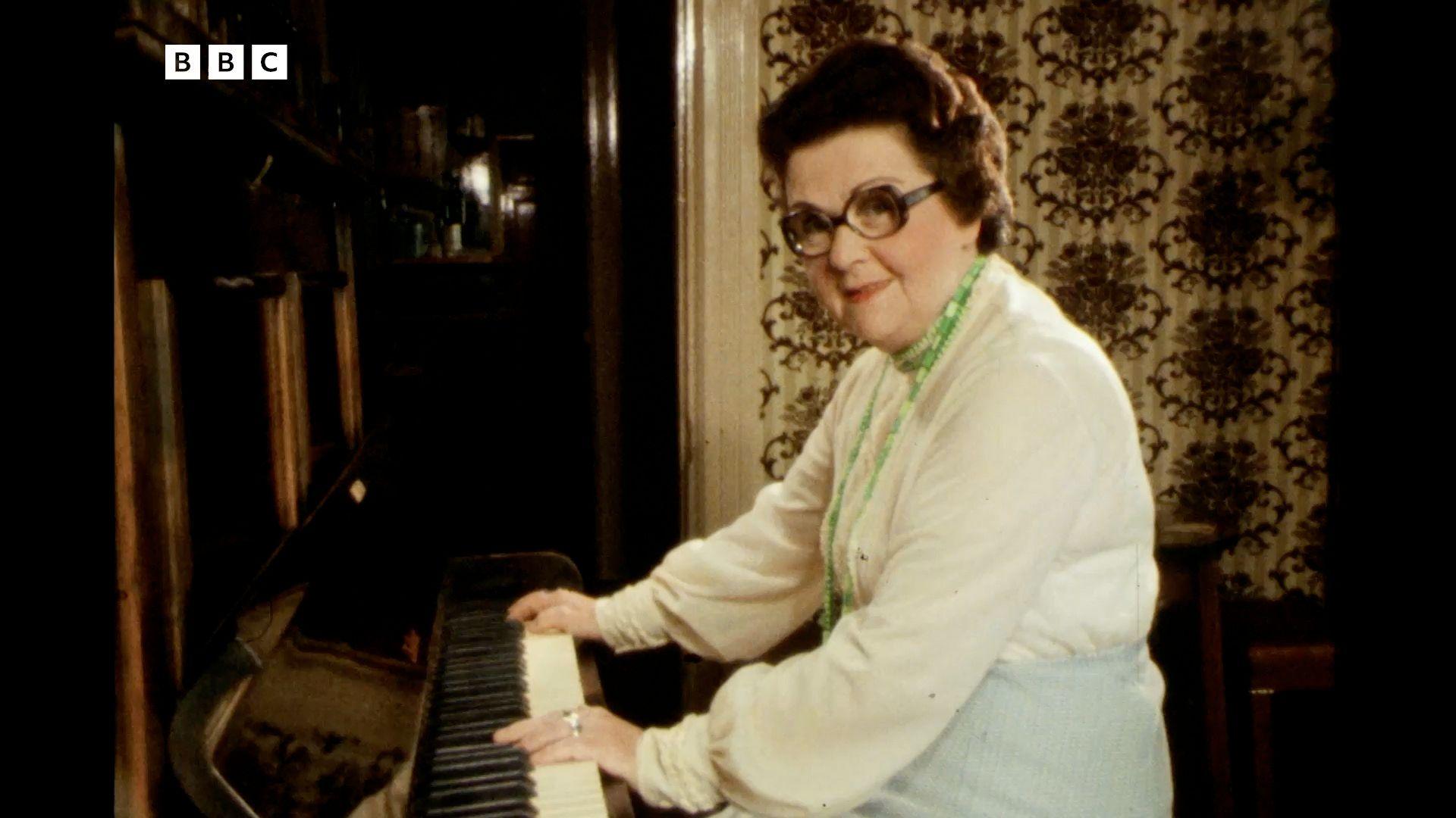 A screenshot from the BBC Look North clip that shows Peggy Fullerton playing the piano while looking into the camera. She's got short, curly brown hair and large frame glasses. She's wearing a white shirt with green beaded necklace. Behind her is a dark brown door and patterned wallpaper.
