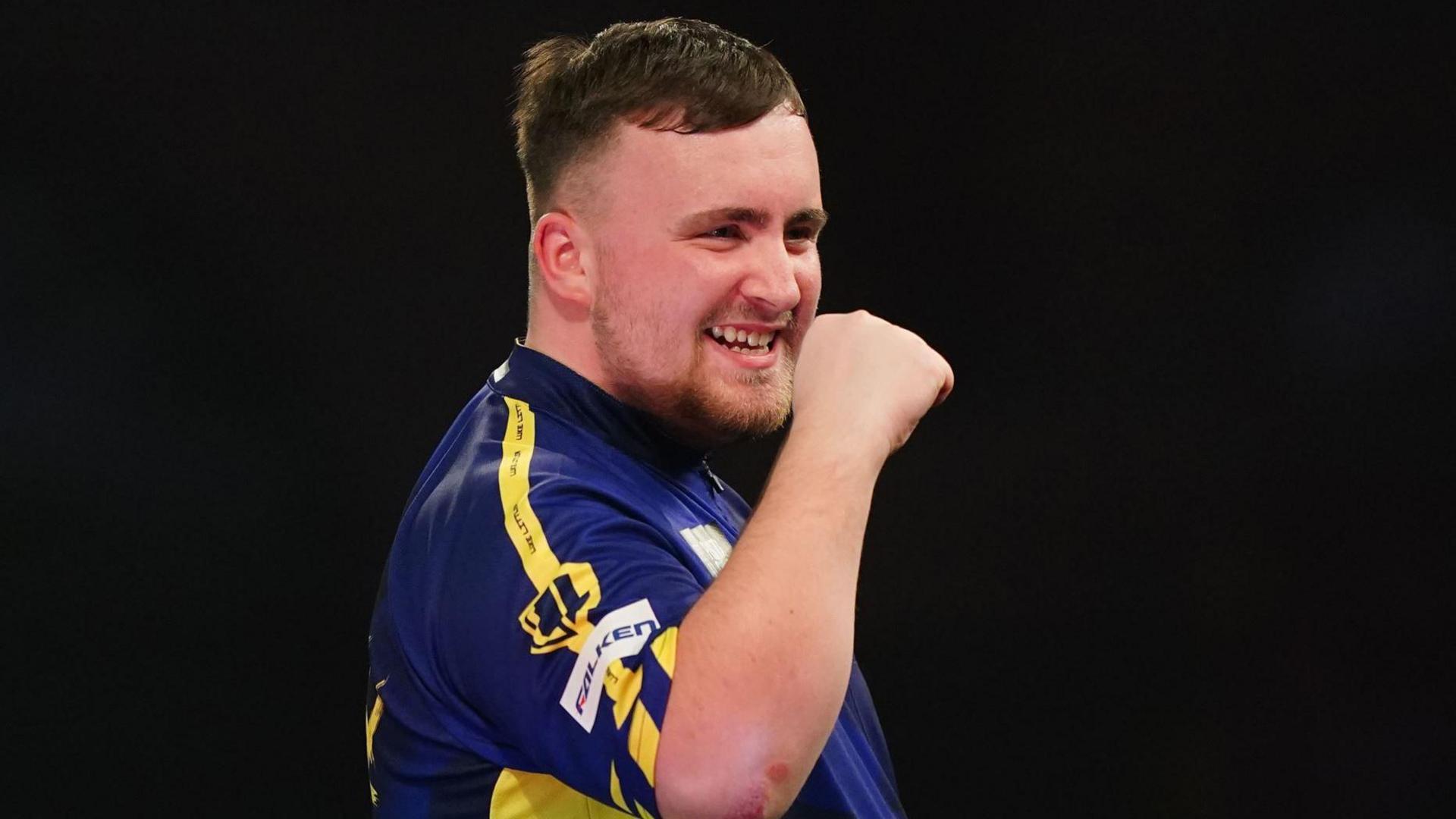 Luke Littler has short brown hair and is wearing a blue and yellow top. He has a clenched fist and is cheering during a darts match.