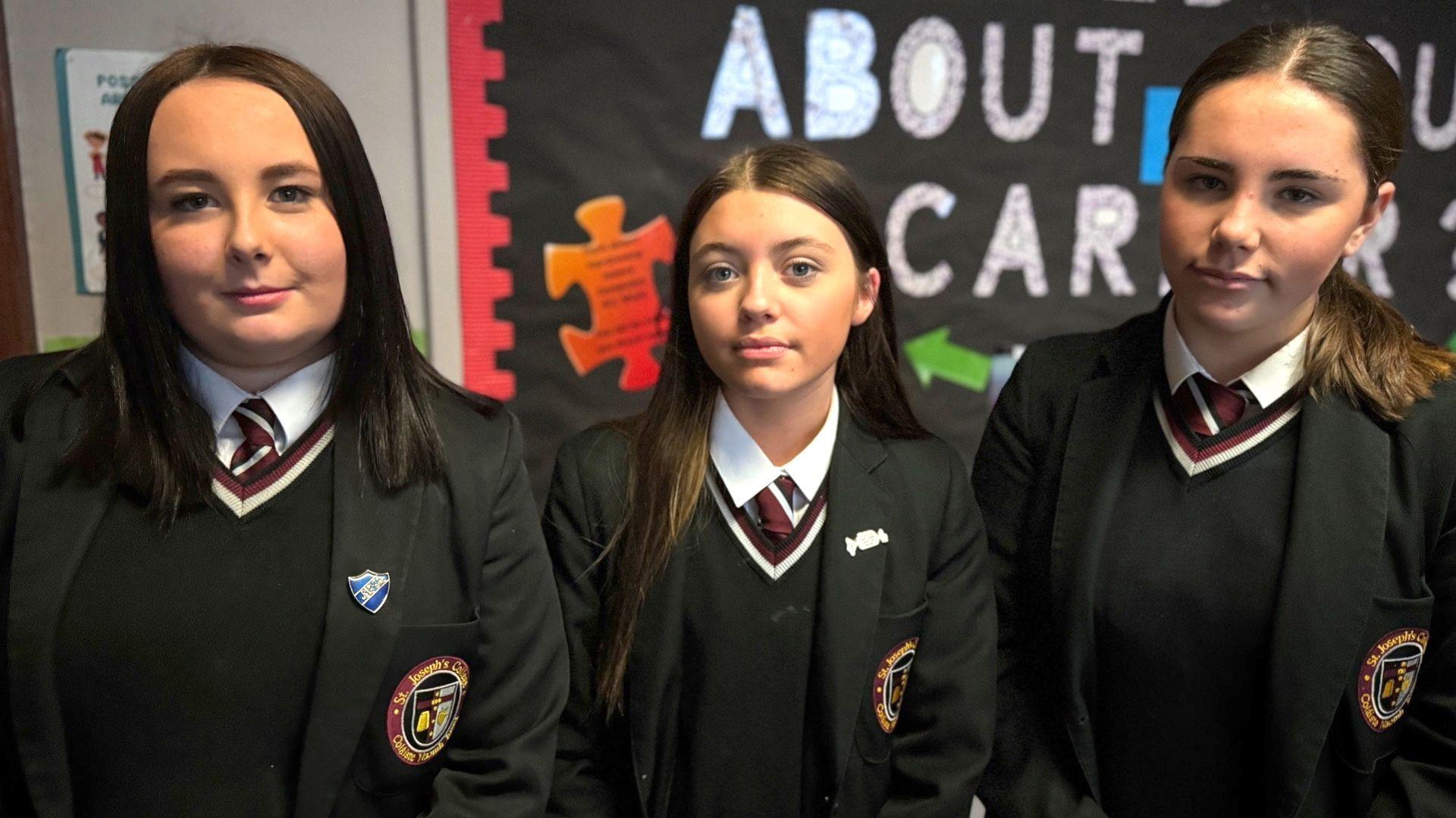 Pictured from left to right is: Cassie, Taigan and Eimear. Cassie has shoulder length black hair, Taigan has long brown hair and Eimear has a dark ponytail. All of the girls are wearing a black blazer which consists of a black blazer, black v-neck jumper, striped burgundy and white tie, and a white shirt.