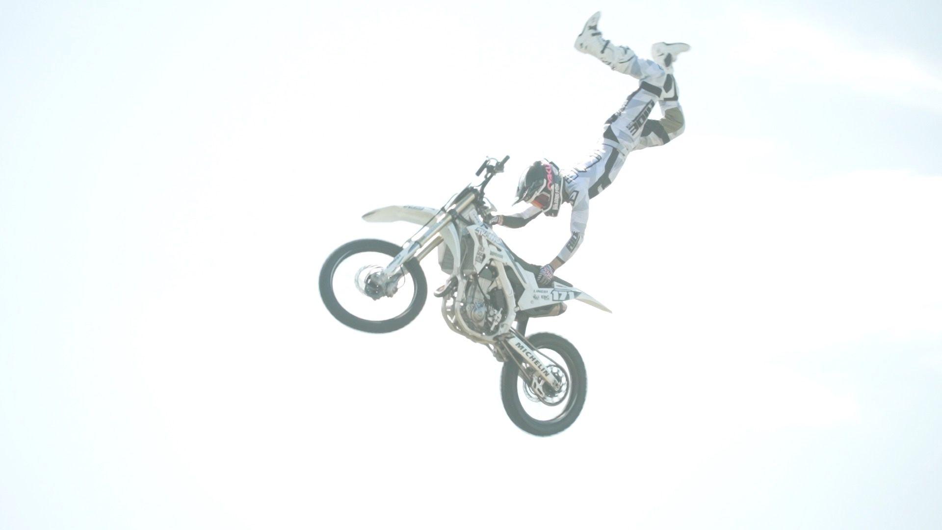 A motorbike rider flips off his bike mid-air during a stunt. He is wearing white overalls and a black helmet. 