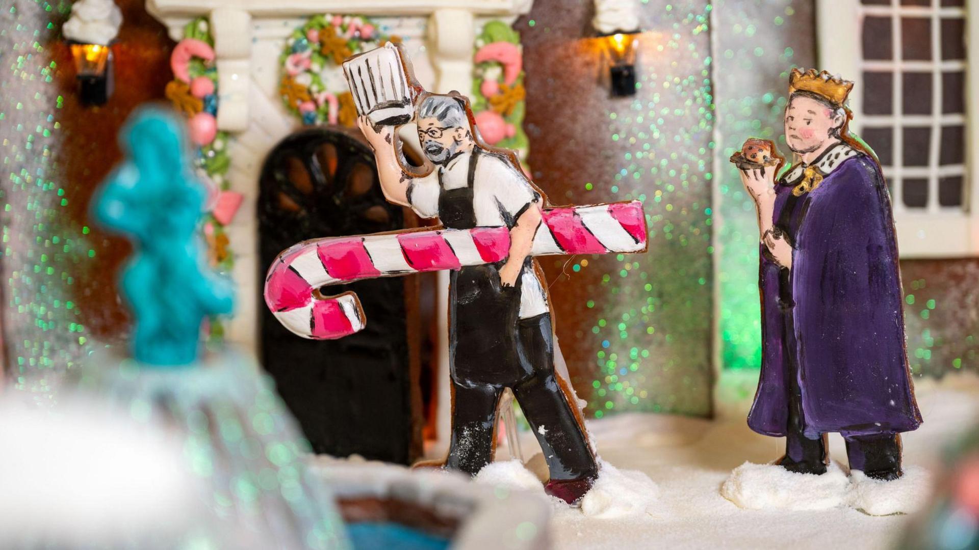 An iced biscuit model of Richard Hughes holding his chef's hat aloft and carrying a red and white striped candy cane is placed outside the gingerbread house. An iced biscuit model of a male character wearing a king's purple velvet cape follows him.