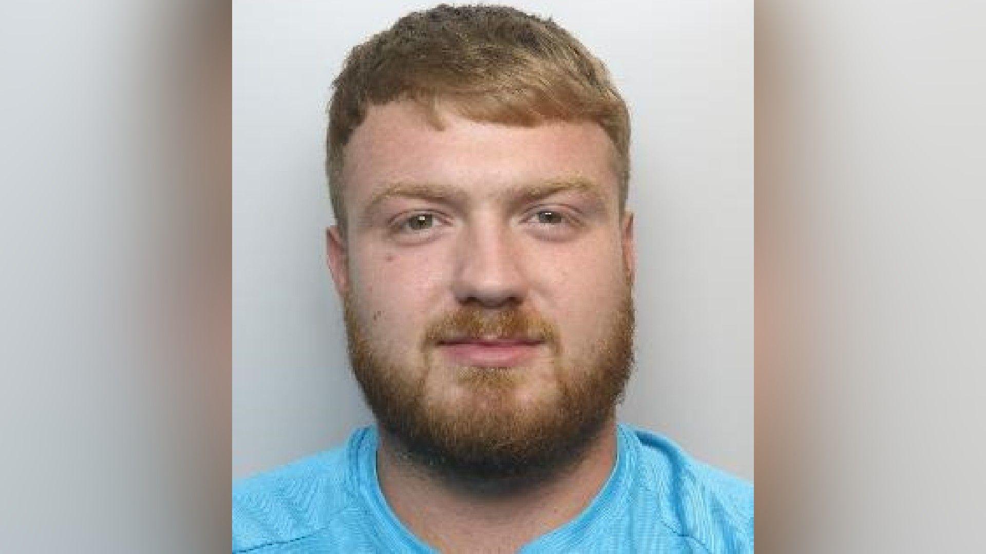 A mugshot of Kaine Hicks, who has strawberry blond hair and a full beard of the same colour. He is smiling at the camera.